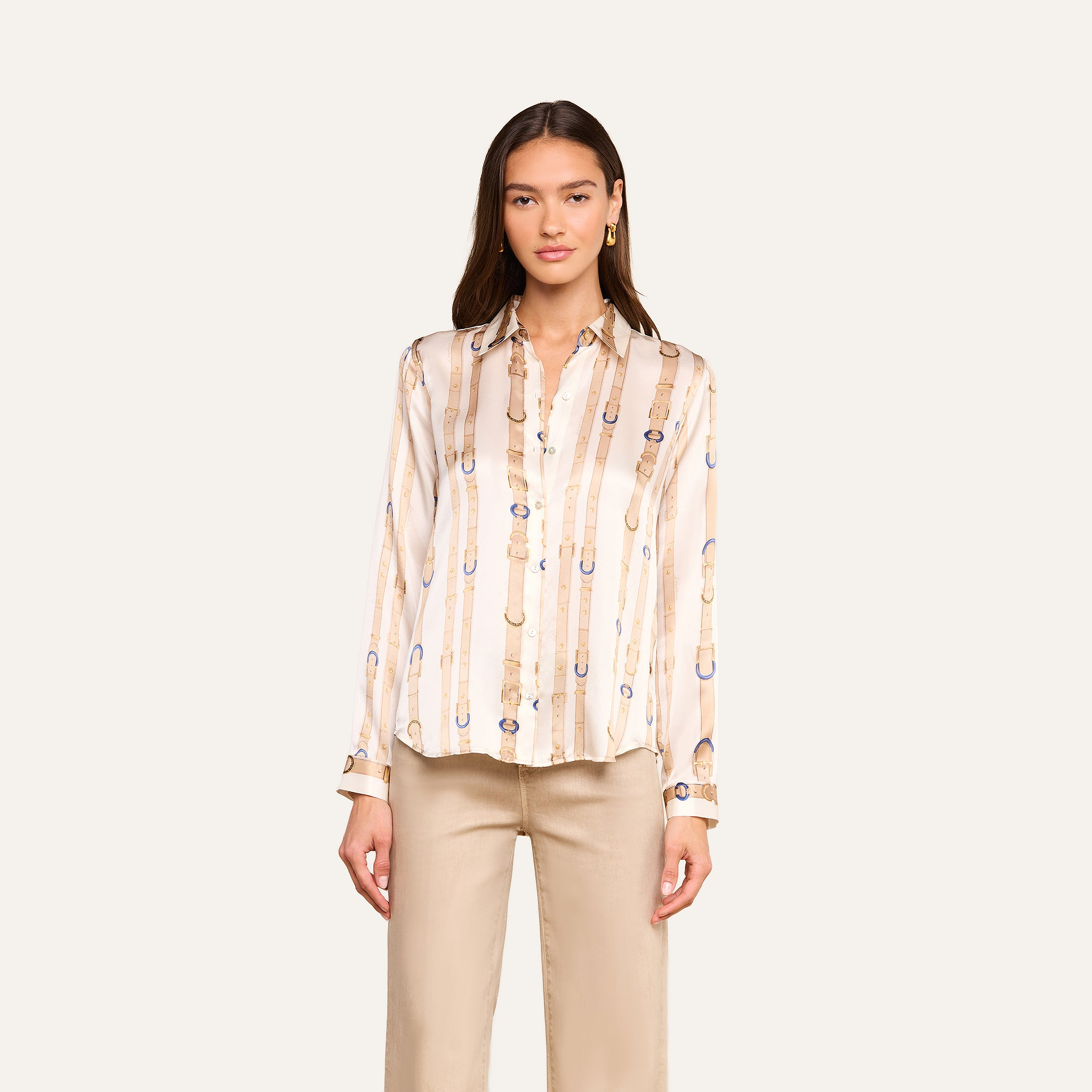 A woman wearing the L’AGENCE Tyler Silk Blouse in a cream color with a belt and buckle pattern, paired with beige pants, stands facing forward against a plain light background.