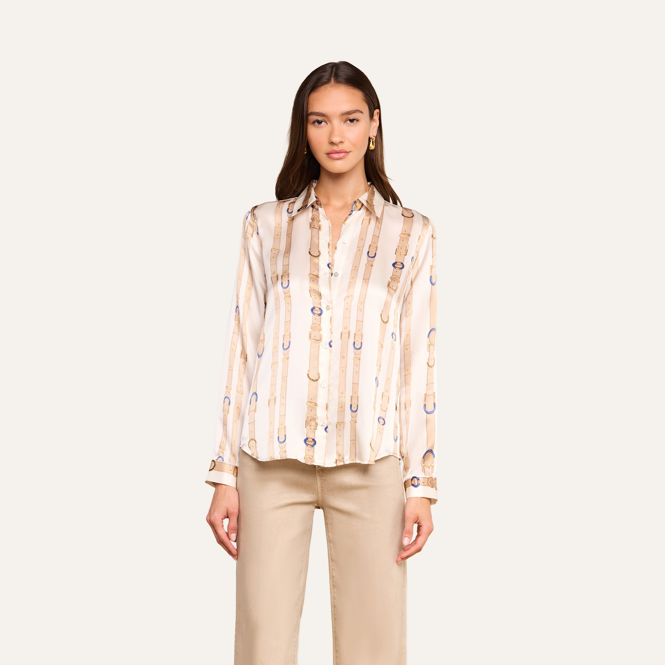 A woman wearing the L’AGENCE Tyler Silk Blouse in a cream color with a belt and buckle pattern, paired with beige pants, stands facing forward against a plain light background.