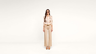 Woman wearing sunglasses, a patterned blouse, beige wide-leg pants, and heels stands facing forward against a plain white background.
