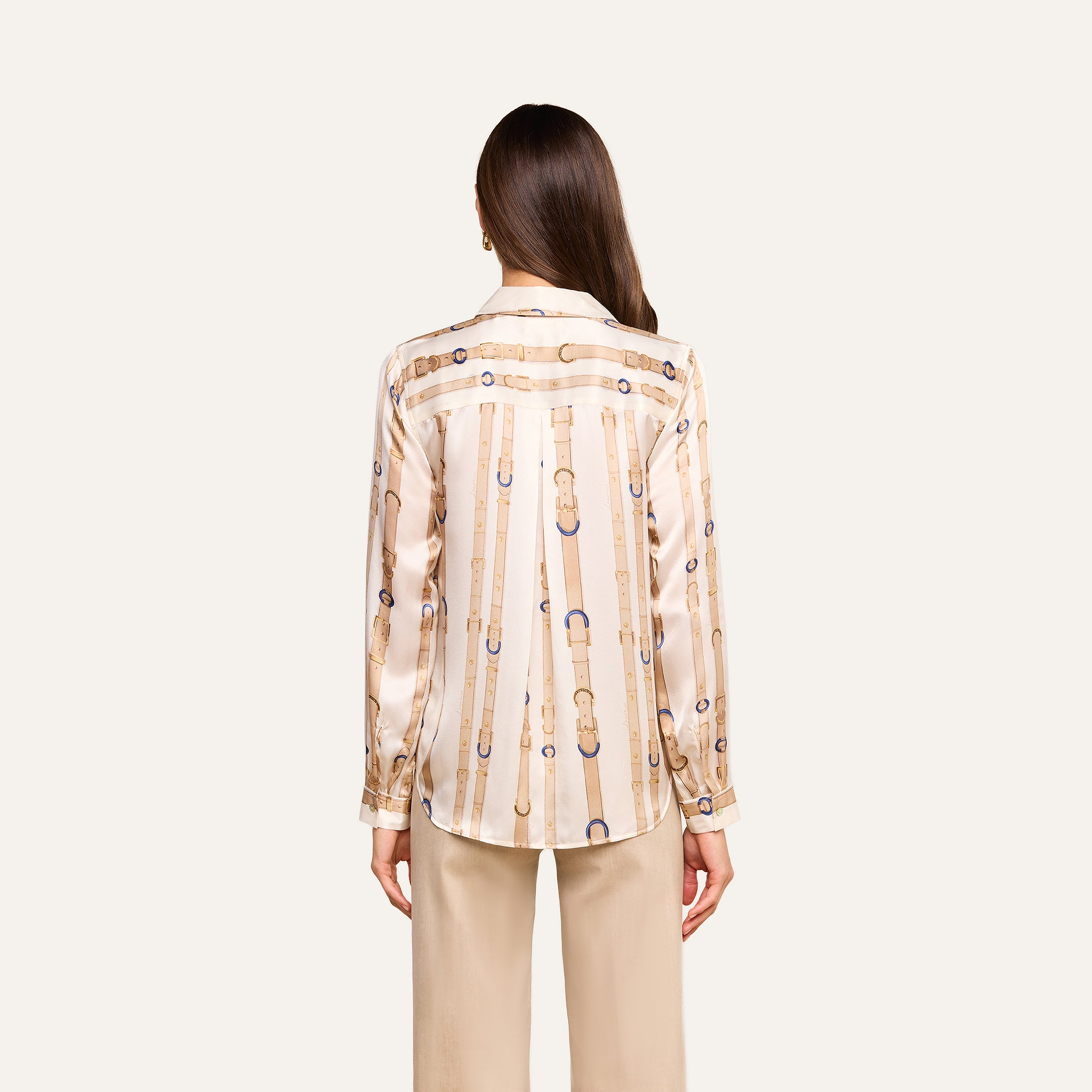 A woman stands with her back to the camera, wearing the Tyler Silk Blouse—a cream-colored, long-sleeve silk button-down featuring a gold and blue chain pattern—paired with beige pants against a plain light background.