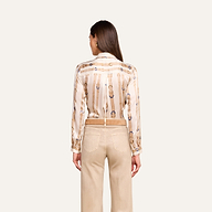 A woman stands facing away, wearing the L’AGENCE Tyler Silk Blouse—long-sleeved with chain designs—and beige pants. Her straight brown hair falls down her back, and her hands rest at her sides against a plain light background.