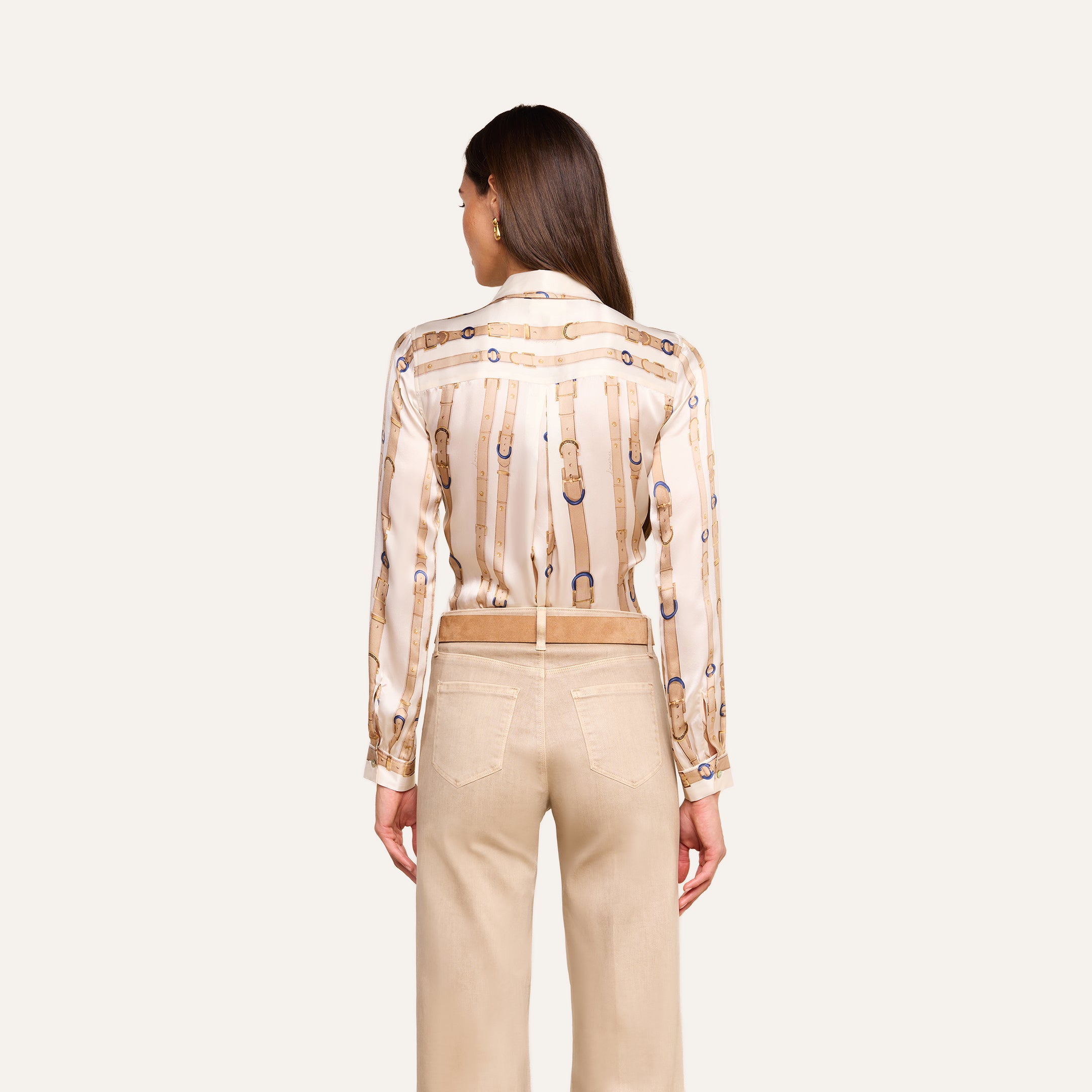 A woman stands facing away, wearing the L’AGENCE Tyler Silk Blouse—long-sleeved with chain designs—and beige pants. Her straight brown hair falls down her back, and her hands rest at her sides against a plain light background.