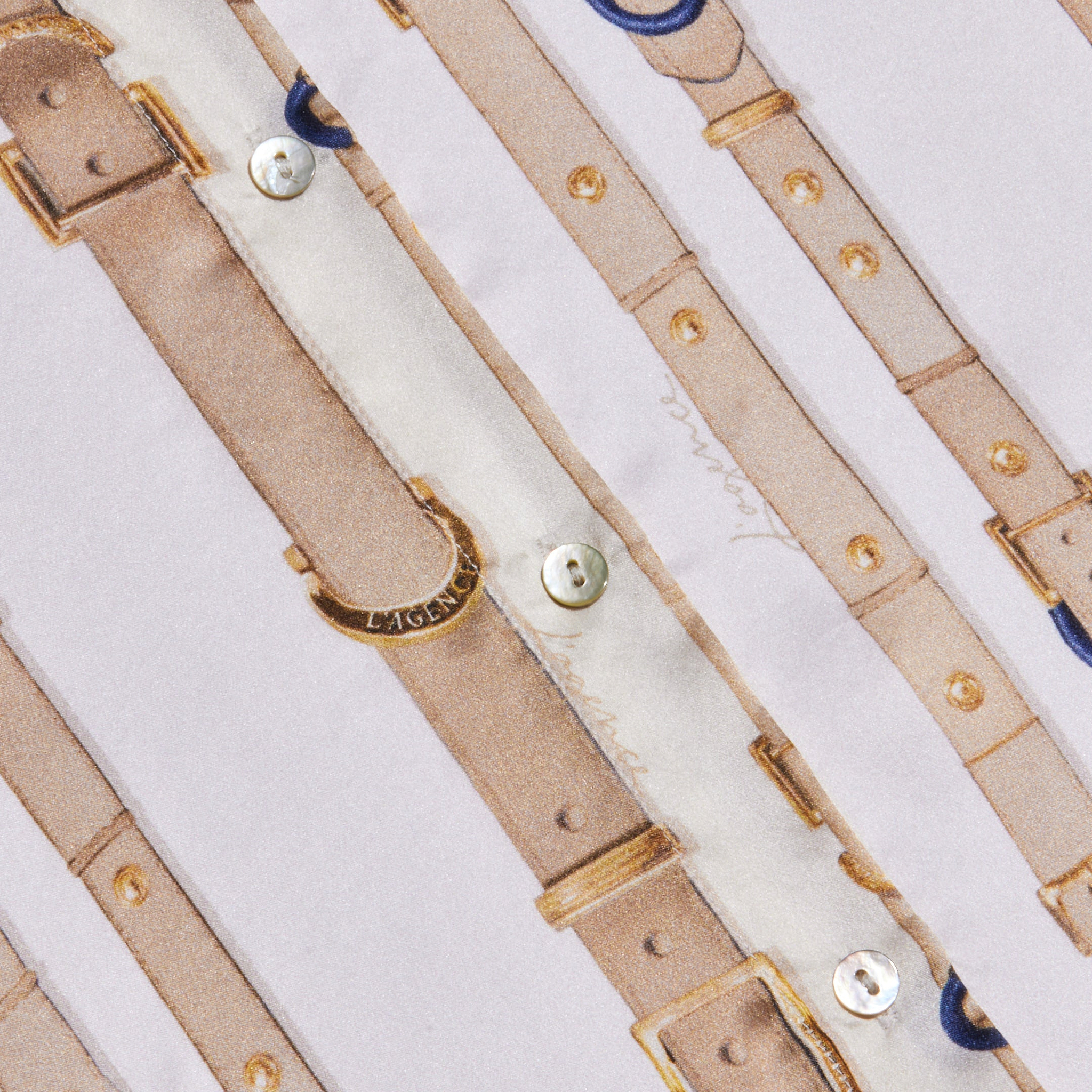 A close-up of fabric patterned with beige straps, gold buckles, and buttons on a white background. The text LAGENCE is visible on a gold buckle, evoking the luxurious detail often found in the Tyler Silk Blouse from L’AGENCE.