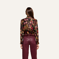 A woman with straight brown hair, worn down, stands against a plain light background, facing away and wearing the L’AGENCE Tyler Silk Blouse featuring a colorful floral pattern paired with burgundy pants.