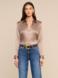 A woman wearing the Tyler Silk Blouse in a shiny, long-sleeve cardamom brown silk, tucked into blue jeans with a brown belt, stands against a plain light background.