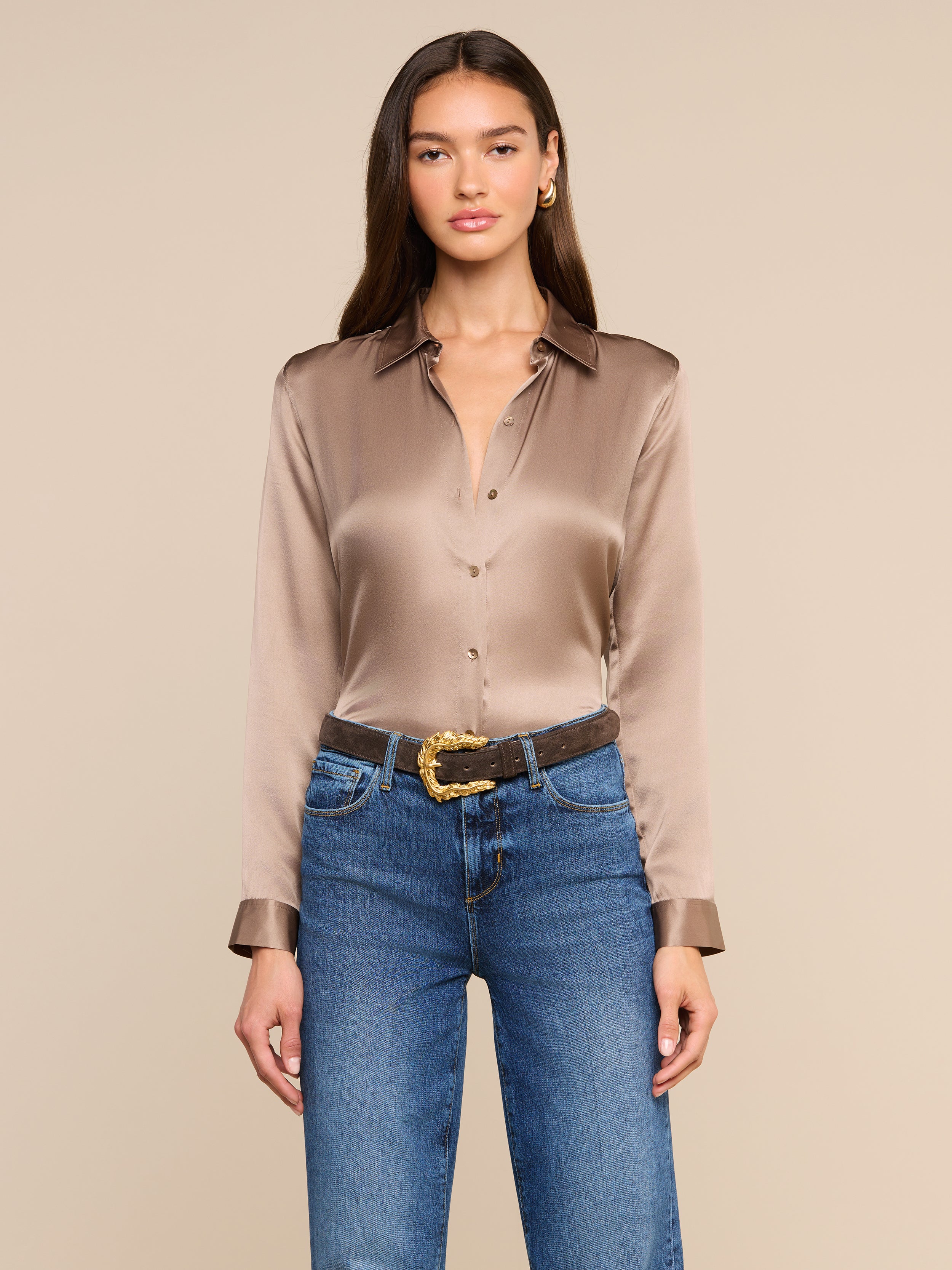 A woman wearing the Tyler Silk Blouse in a shiny, long-sleeve cardamom brown silk, tucked into blue jeans with a brown belt, stands against a plain light background.