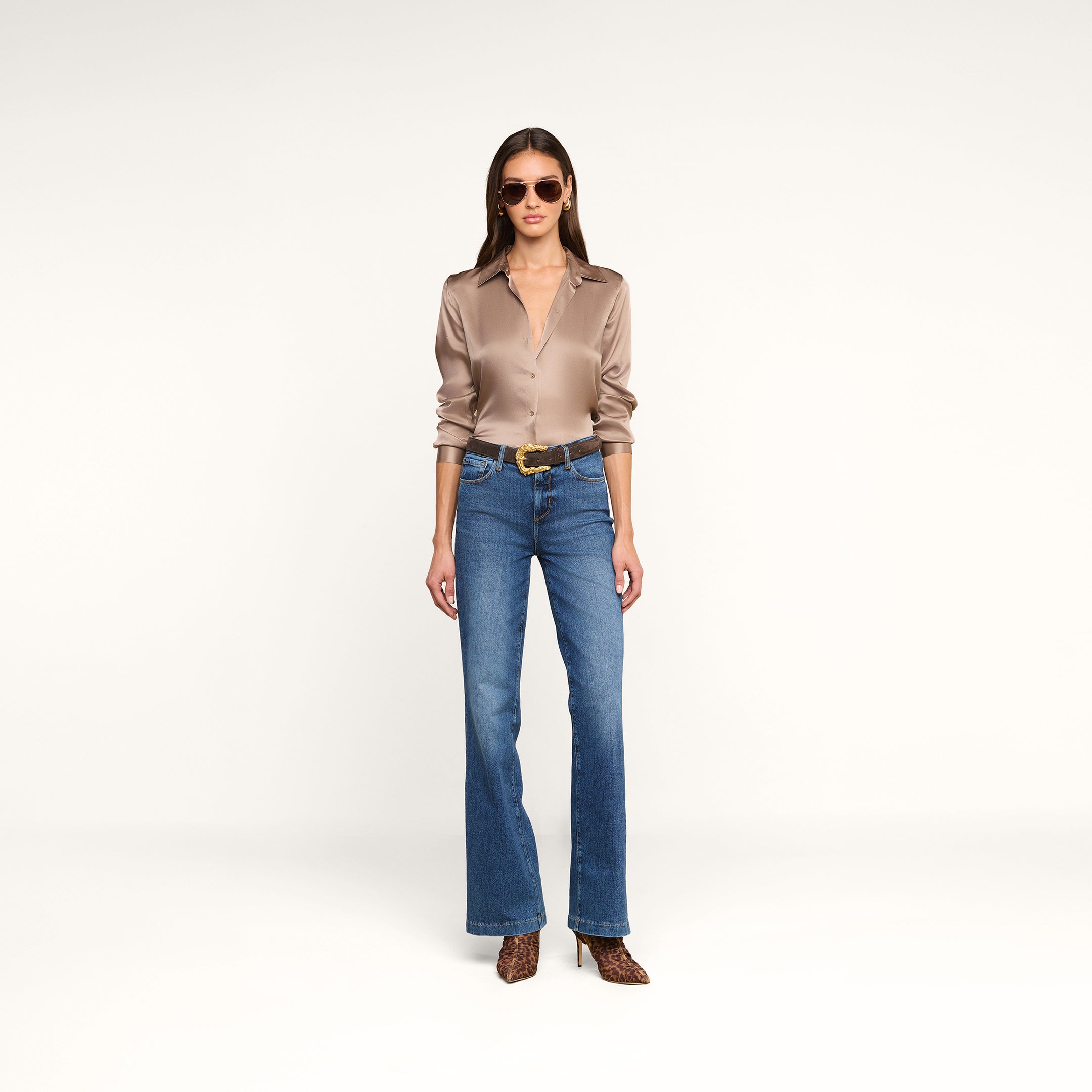 A woman stands against a plain background, wearing sunglasses, the Tyler Silk Blouse—a long-sleeve taupe button-down—blue wide-leg jeans, a black belt with a gold buckle, and leopard print heels. She faces forward with her hands at her sides.