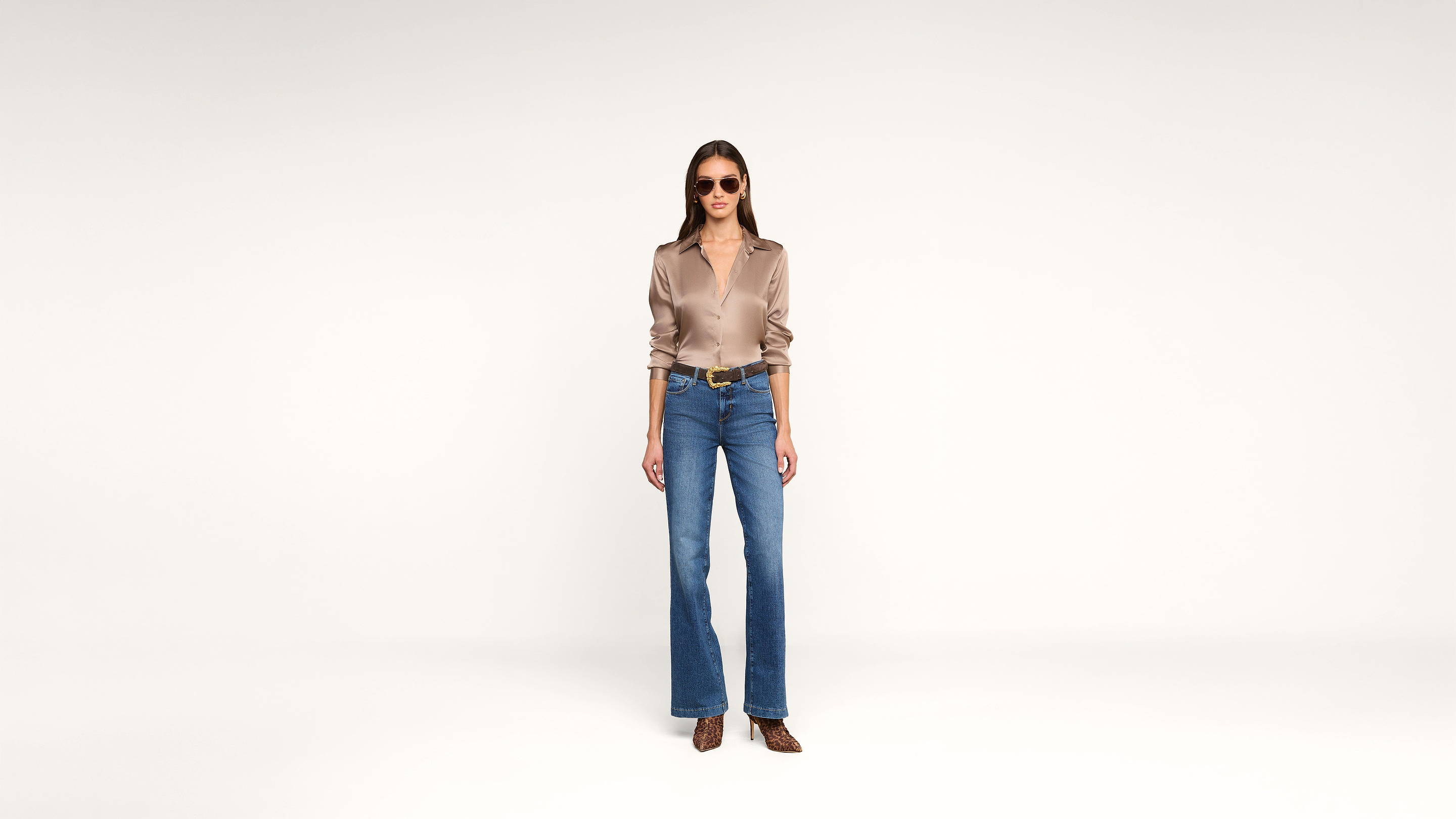 A woman stands against a plain white background, wearing sunglasses, a silky beige blouse, blue wide-leg jeans, a brown belt, and brown pointed shoes. She faces forward with a relaxed posture.
