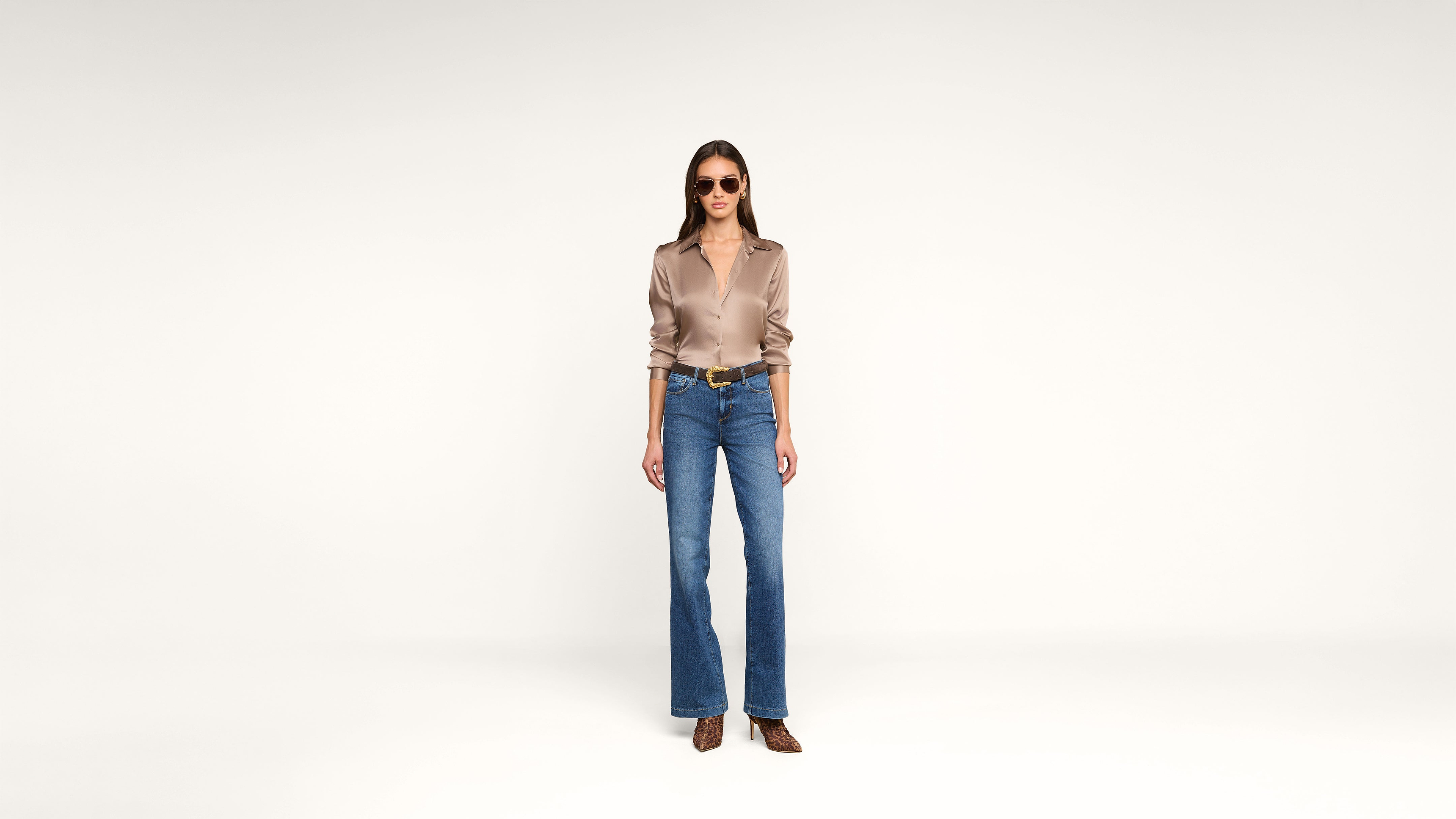 A woman stands against a plain white background, wearing sunglasses, a silky beige blouse, blue wide-leg jeans, a brown belt, and brown pointed shoes. She faces forward with a relaxed posture.