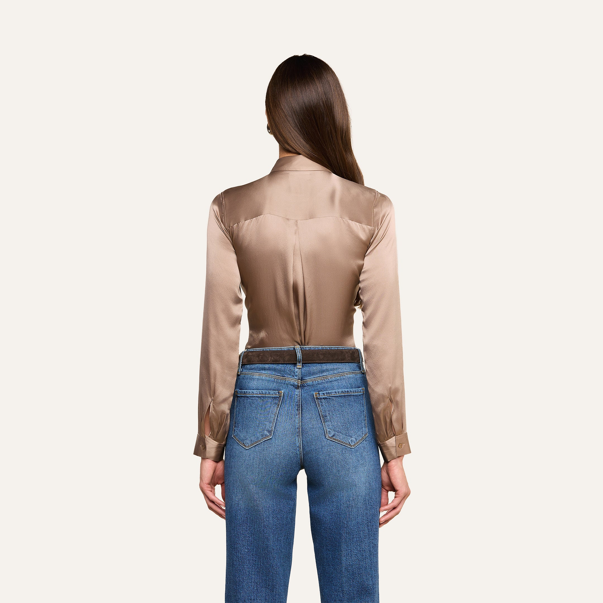 A woman with long brown hair, seen from behind, wears the Tyler Silk Blouse in cardamom brown with long sleeves and blue jeans against a plain light background.