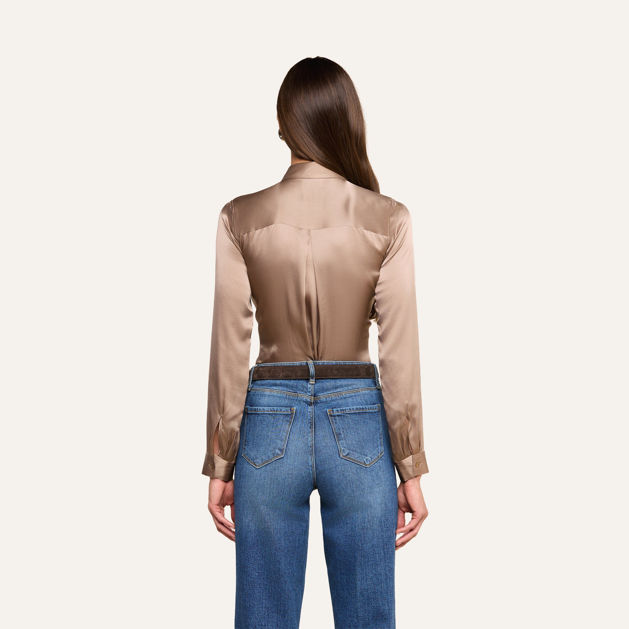 A woman with long brown hair, seen from behind, wears the Tyler Silk Blouse in cardamom brown with long sleeves and blue jeans against a plain light background.