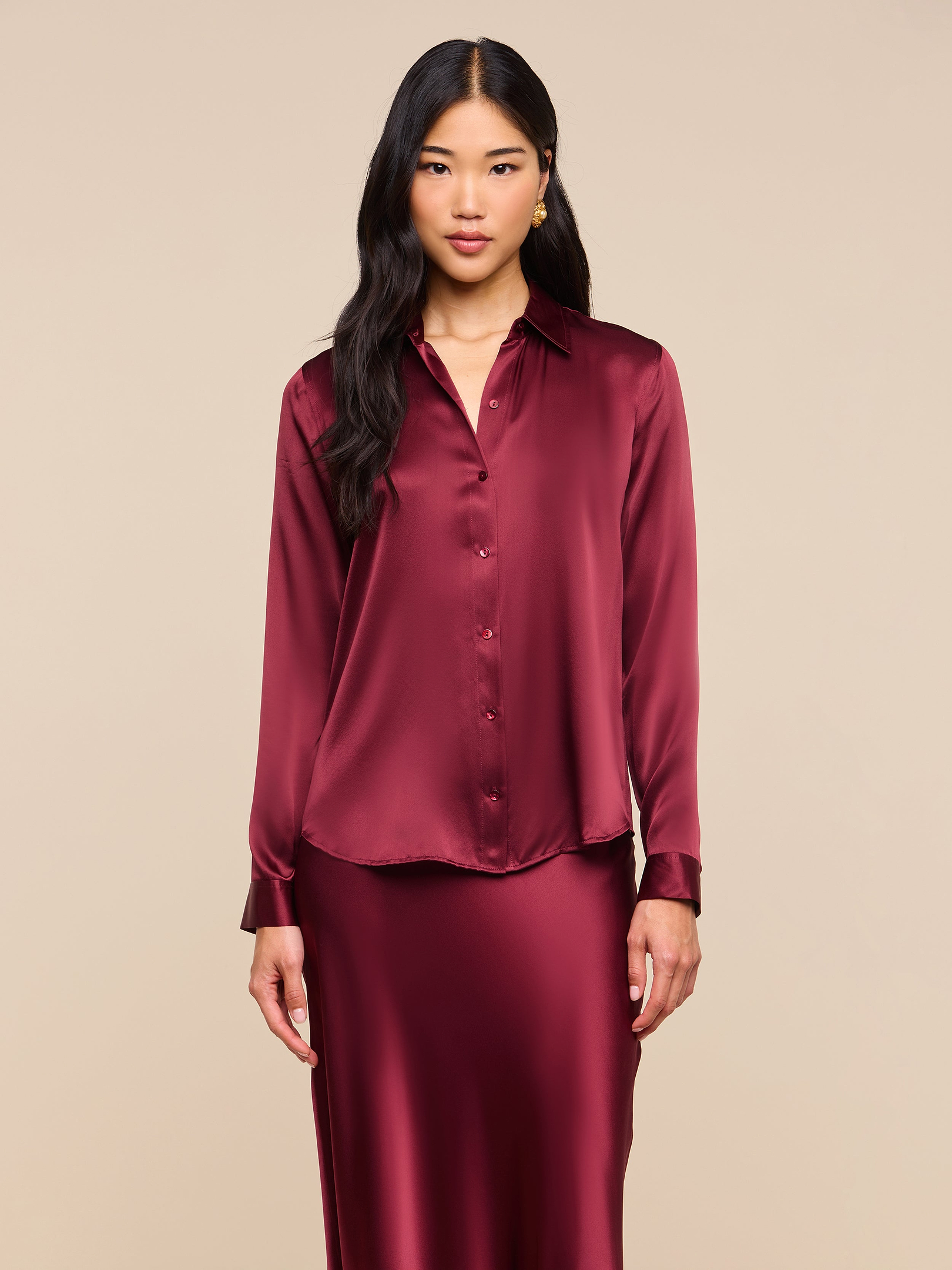 A woman wearing the Tyler Silk Blouse—a long-sleeved, button-up silk charmeuse top in merlot red—and a matching skirt, stands against a plain light background, looking at the camera.