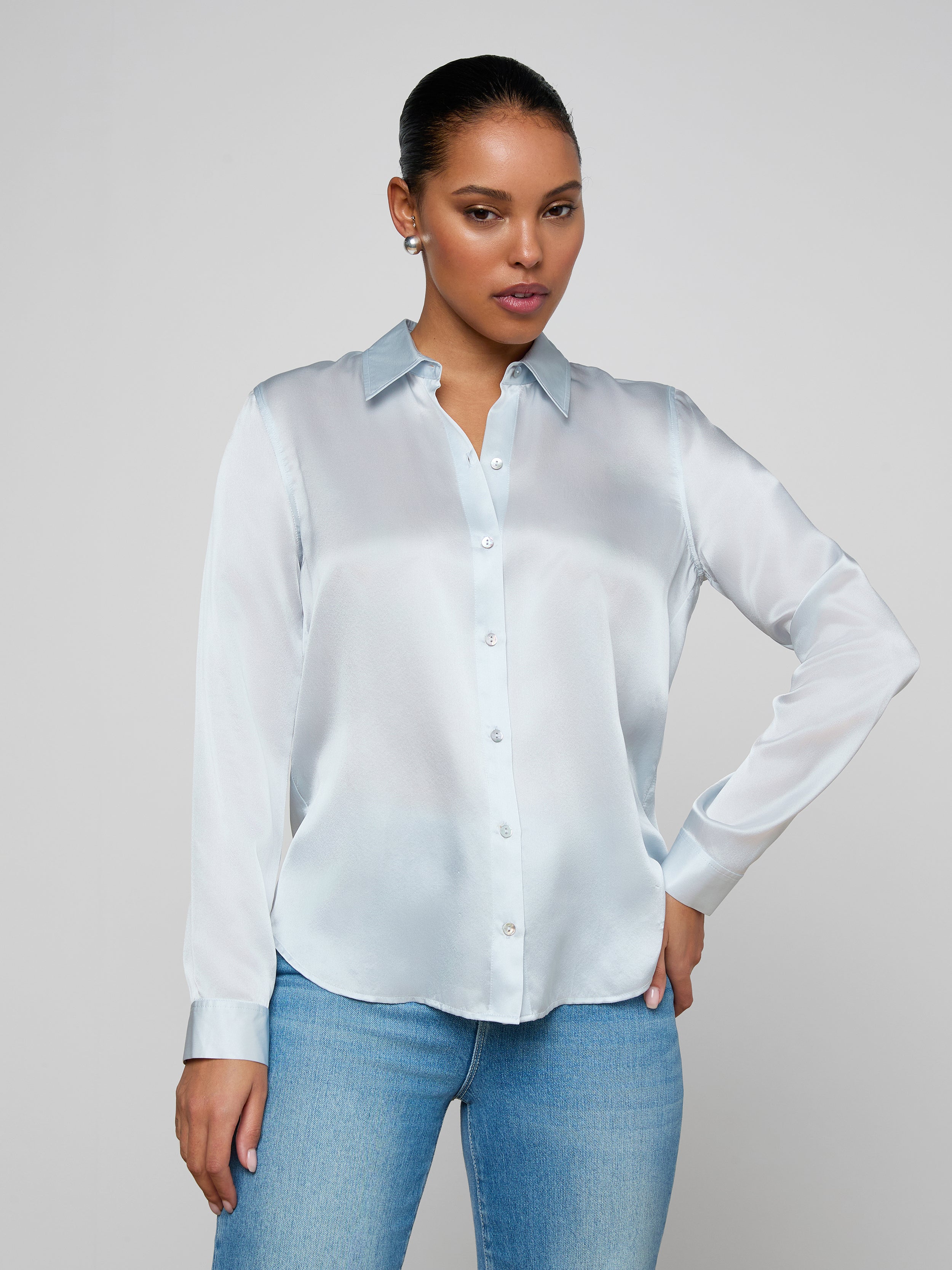 Wearing the Tyler Silk Blouse in pale mist blue and a pair of blue jeans, a person poses against a plain light background with one hand on their hip and the other relaxed by their side.