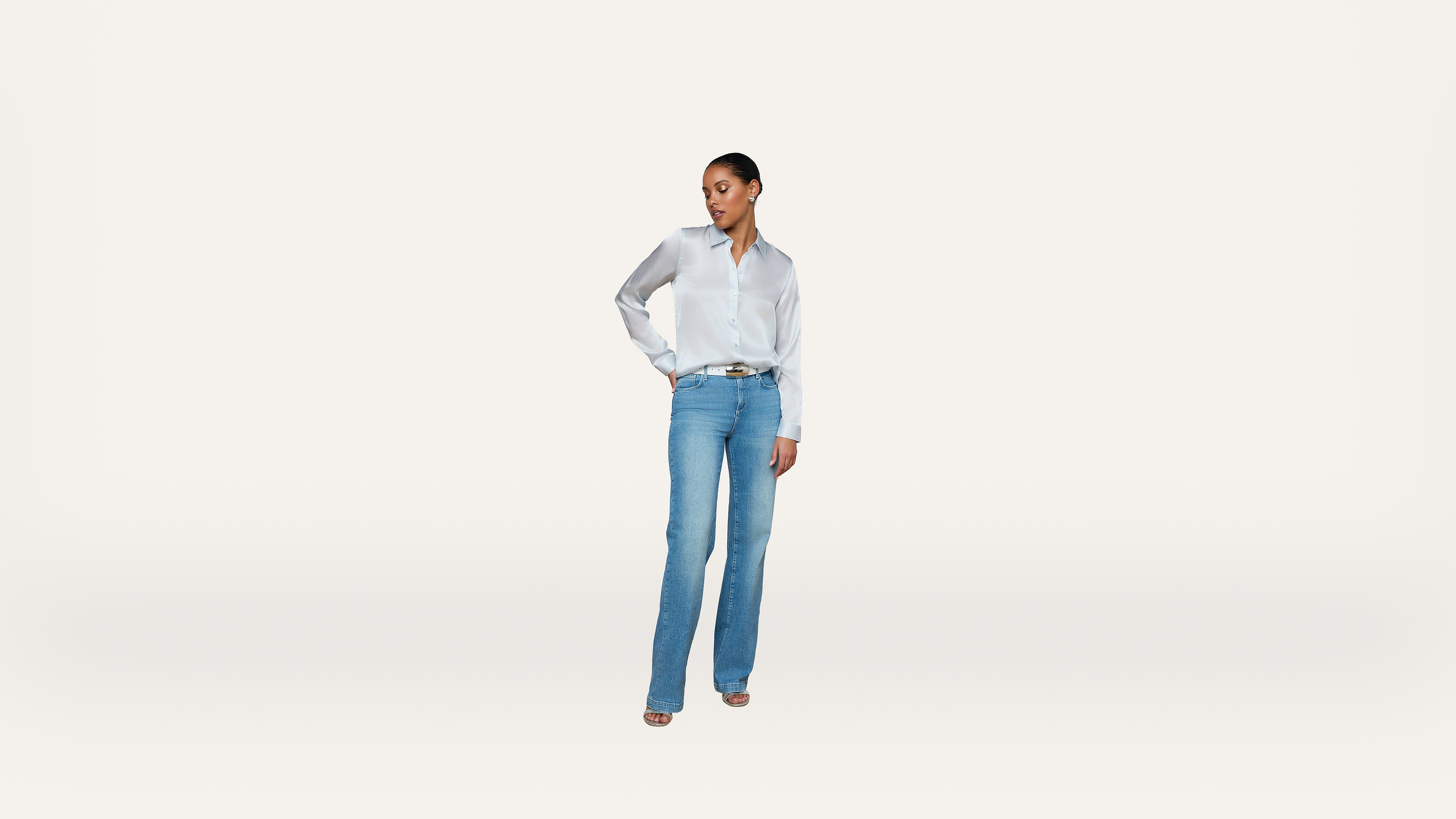 A woman stands against a white background, wearing a white button-down shirt, blue jeans, and heels. She has one hand on her hip and looks to the side.