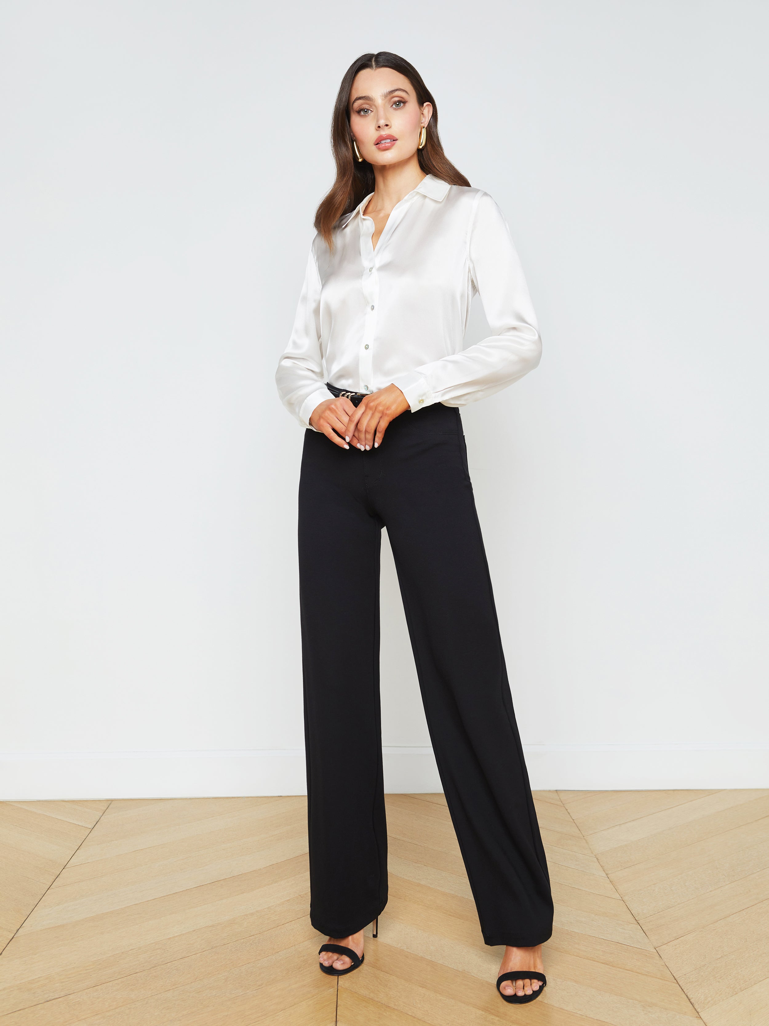 A woman stands against a plain background wearing the ivory Tyler Silk Blouse, black wide-leg pants, and black heeled sandals.