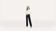 A woman stands against a plain white background, wearing a white long-sleeve blouse, black wide-leg trousers, and black open-toe heels.