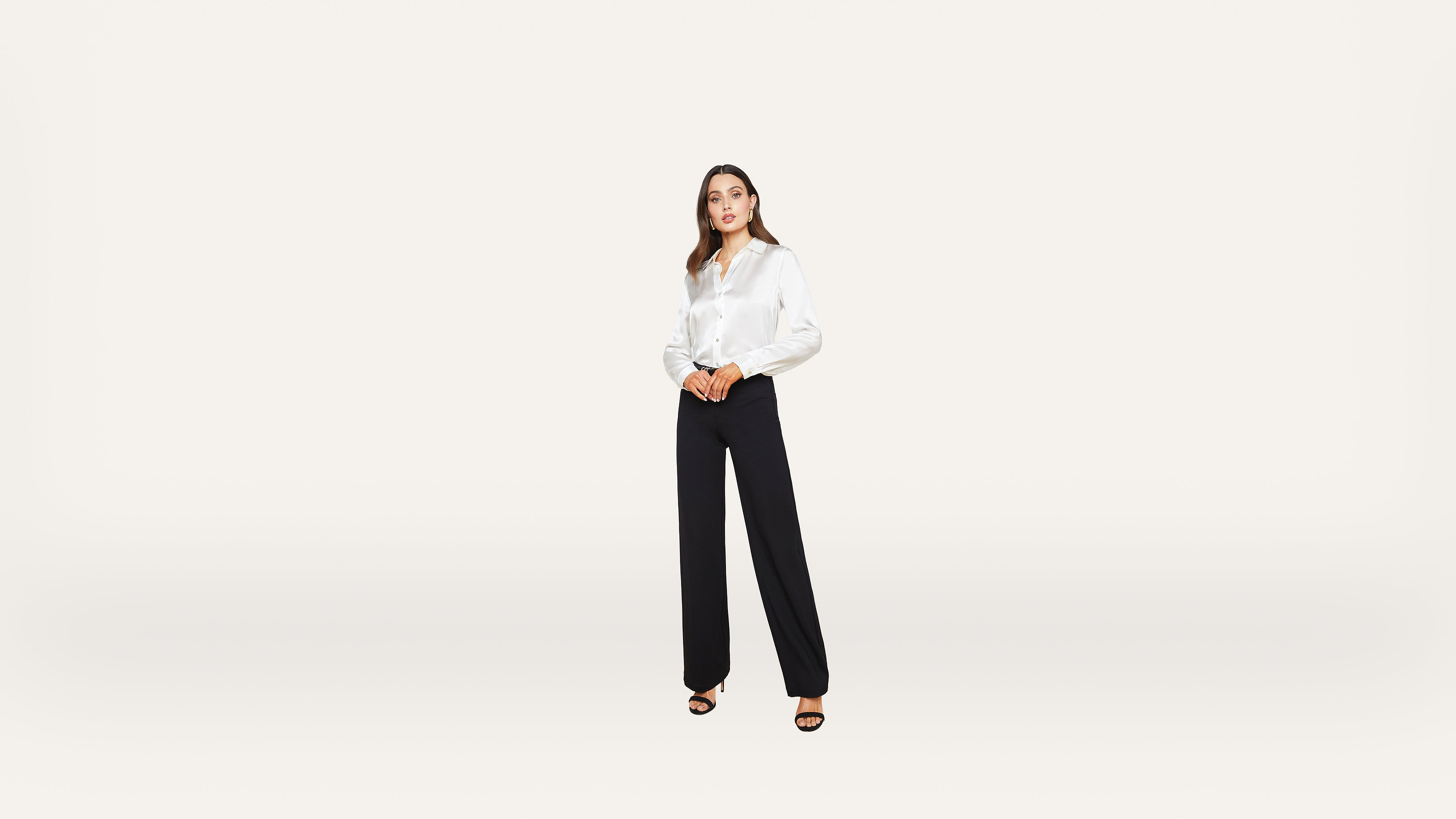 A woman stands against a plain white background, wearing a white long-sleeve blouse, black wide-leg trousers, and black open-toe heels.