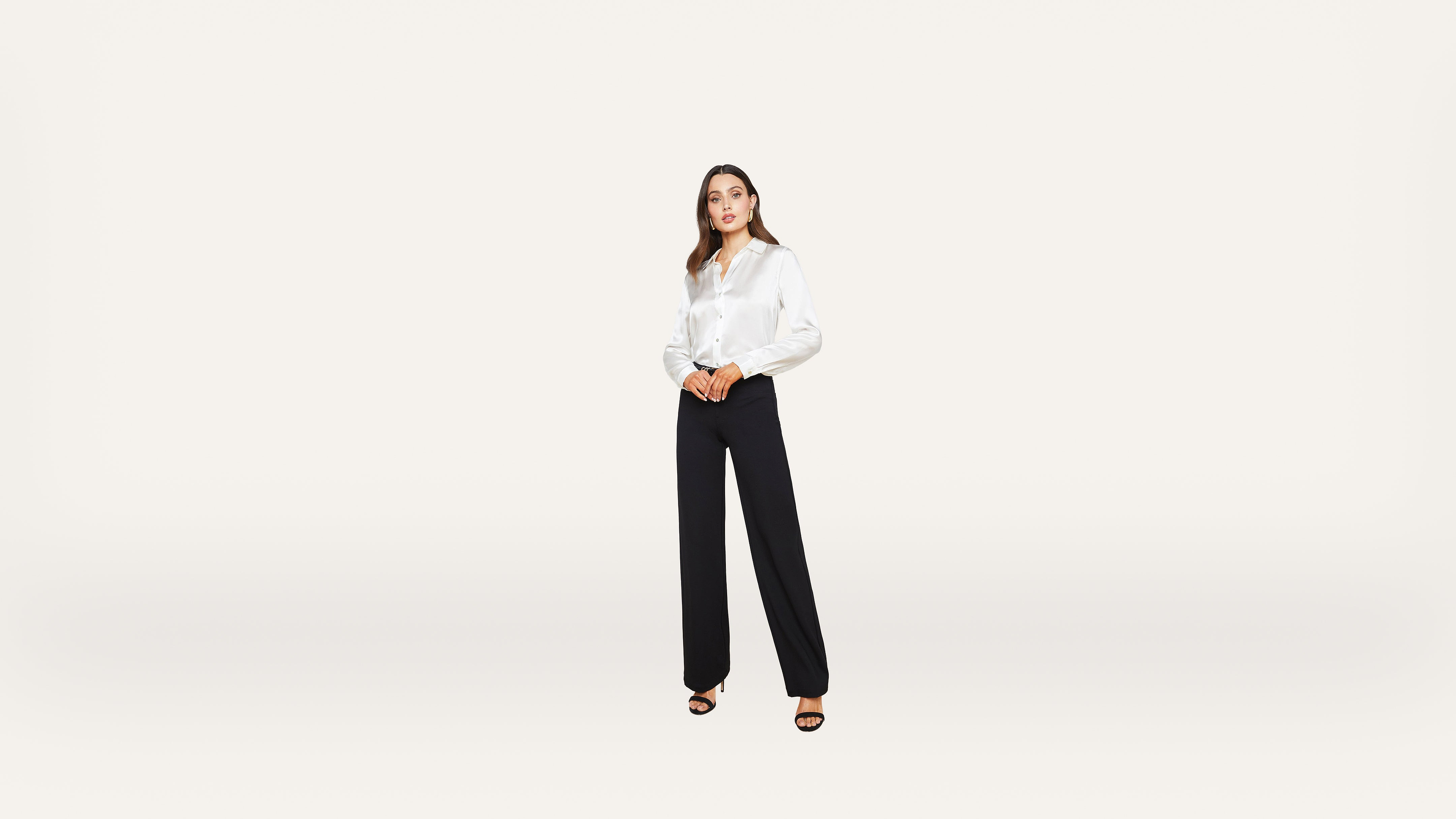 A woman stands against a plain white background, wearing a white long-sleeve blouse, black wide-leg trousers, and black open-toe heels.