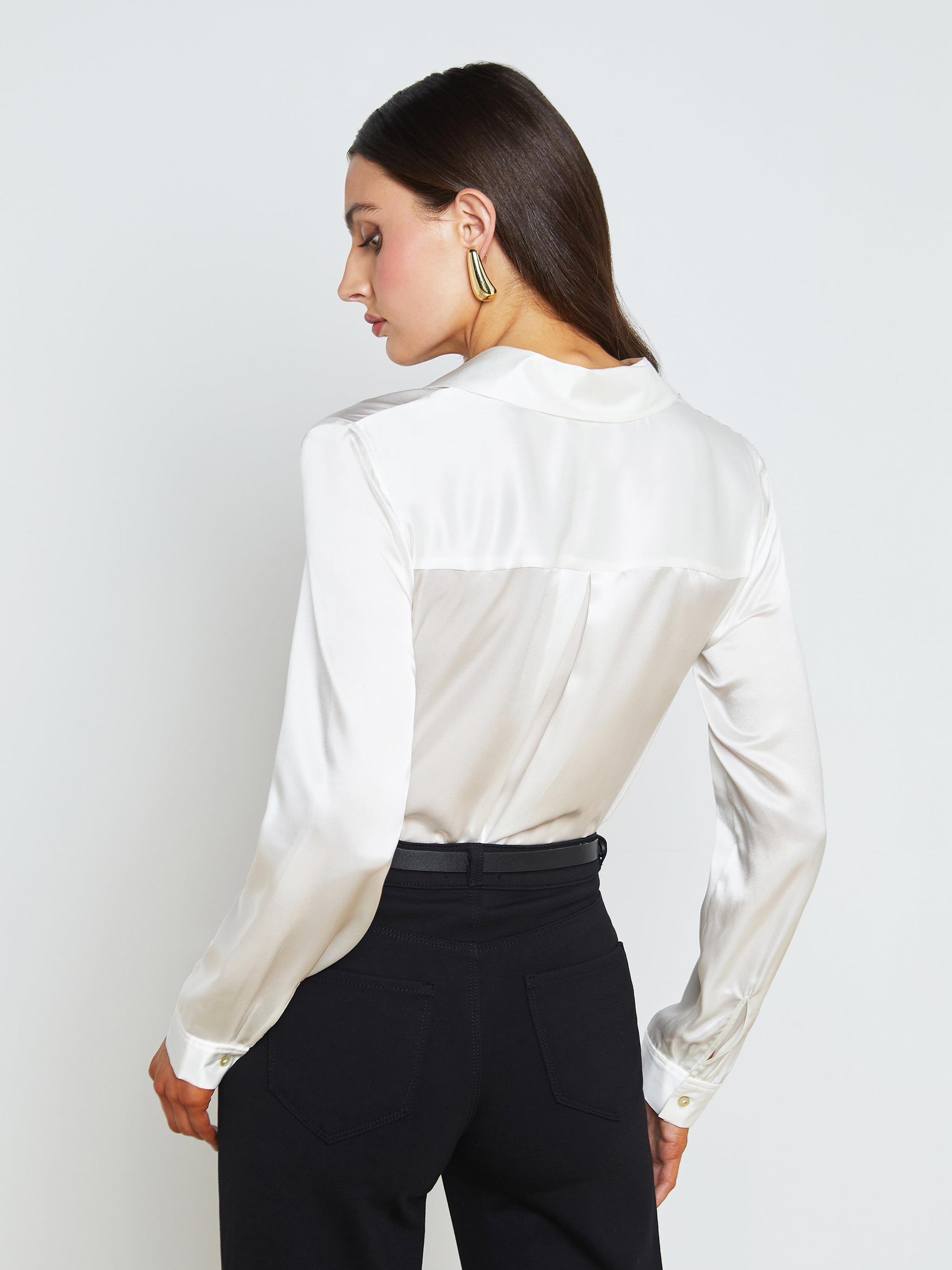 Woman with straight brown hair wearing the Tyler Silk Blouse in ivory and black pants, facing away and slightly turning her head to the side. She is also wearing gold statement earrings against a plain background.
