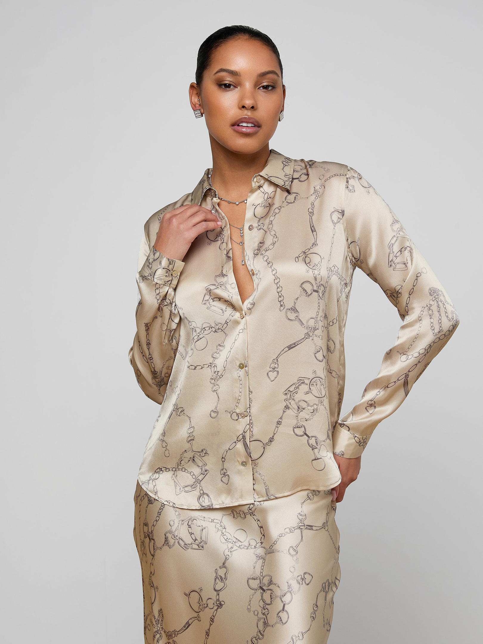 A woman stands confidently against a plain light background, wearing the Tyler Silk Blouse in cream and a matching skirt with a subtle floral pattern; she has one hand on her hip and the other holding her collar.