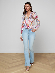 A woman stands confidently against a plain white background, wearing the colorful Tyler Silk Blouse from L’AGENCE paired with light blue flared jeans, her hand in her pocket and open-toed heels.