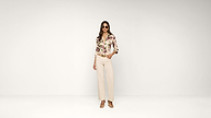 A woman wearing sunglasses, a floral blouse, cream wide-leg pants, and heeled sandals stands confidently against a plain white background.