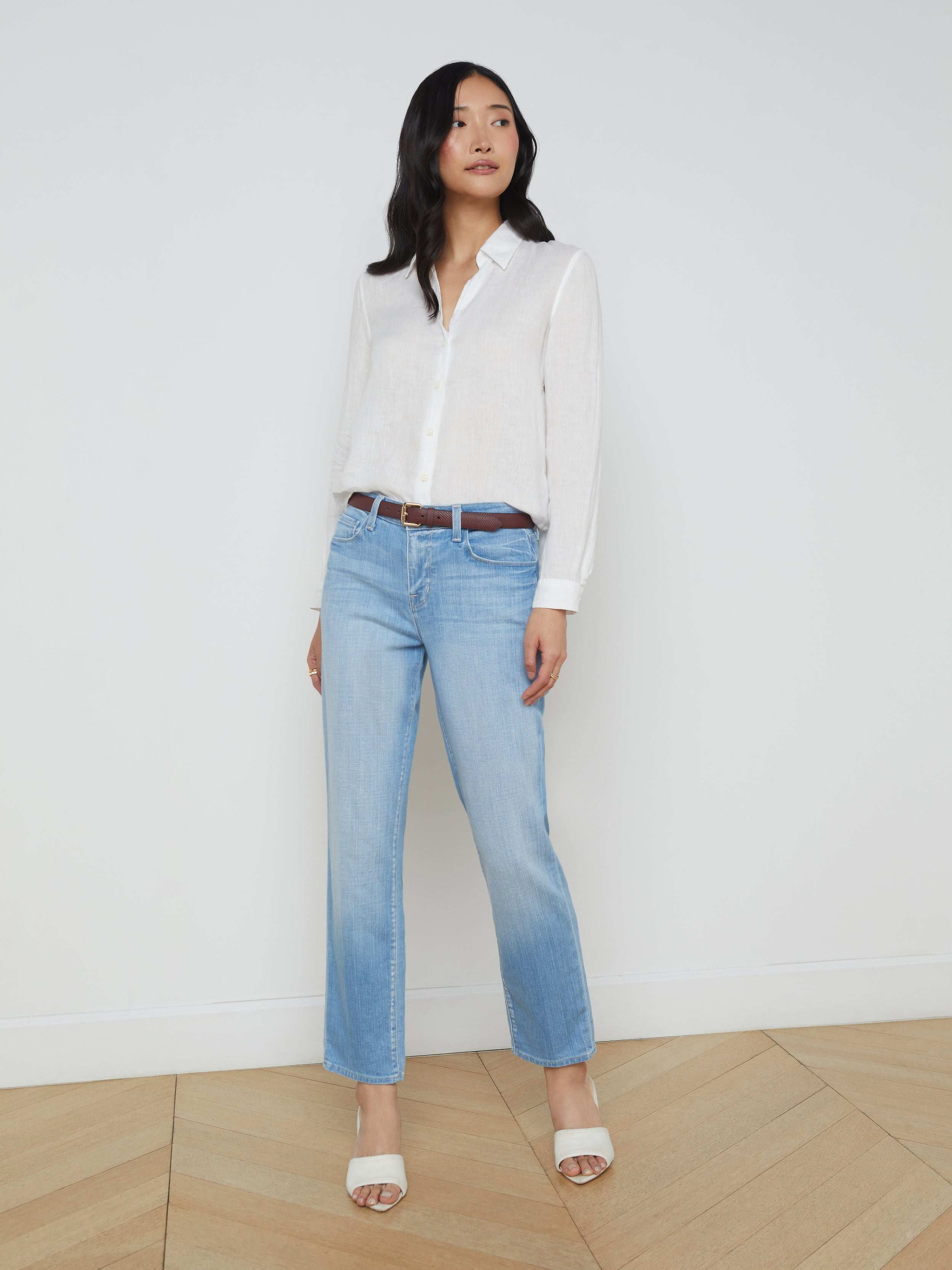 A woman stands against a plain white background, wearing the Tyler Linen Blouse—a white long sleeve button-down summer shirt—paired with light blue jeans, a brown belt, and beige open-toe heels.