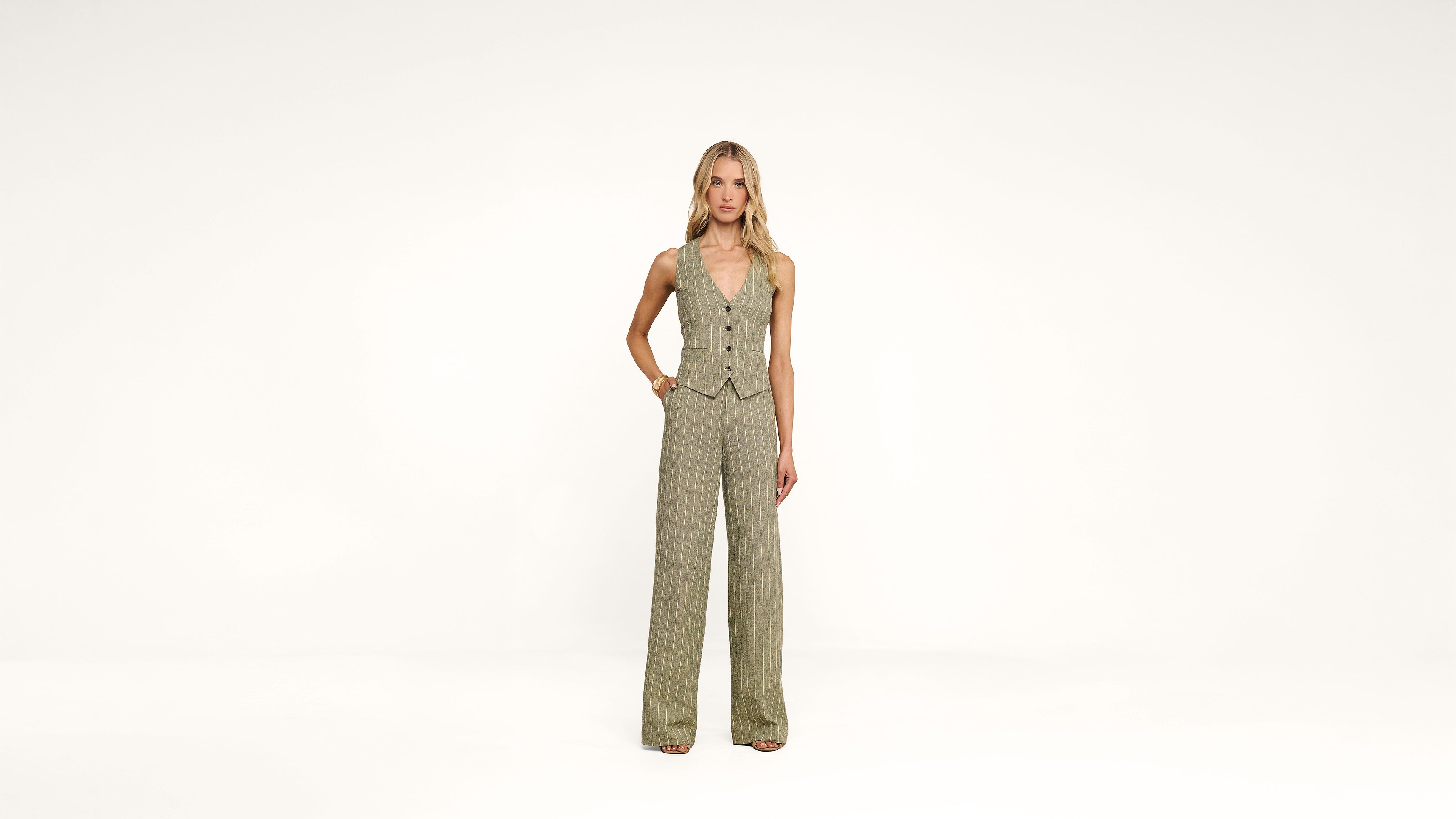 A person stands against a plain white background, wearing a sleeveless beige vest and matching wide-leg pants, with hands in pockets and looking toward the camera.