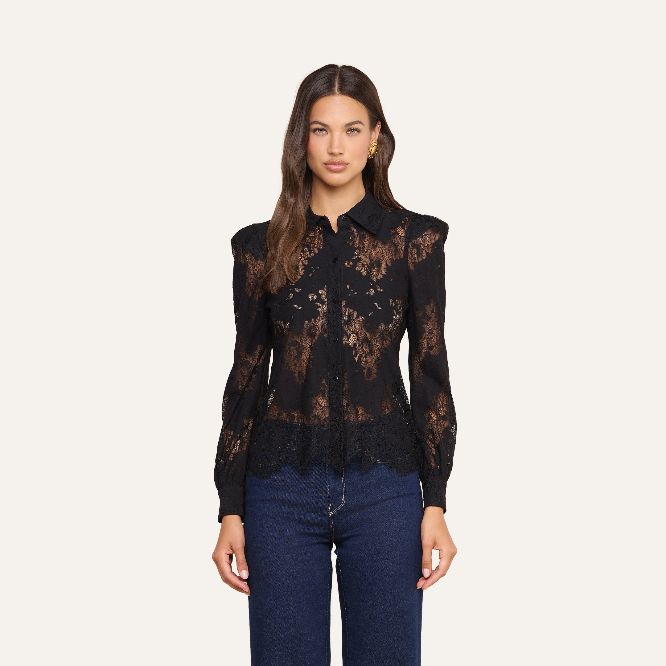 Una donna dai lunghi capelli castani indossa la camicetta Jenica Lace, un modello femminile in pizzo nero con bottoni e maniche lunghe, abbinata a un paio di jeans blu scuro. È in piedi davanti a uno sfondo beige a tinta unita e guarda direttamente verso la fotocamera.