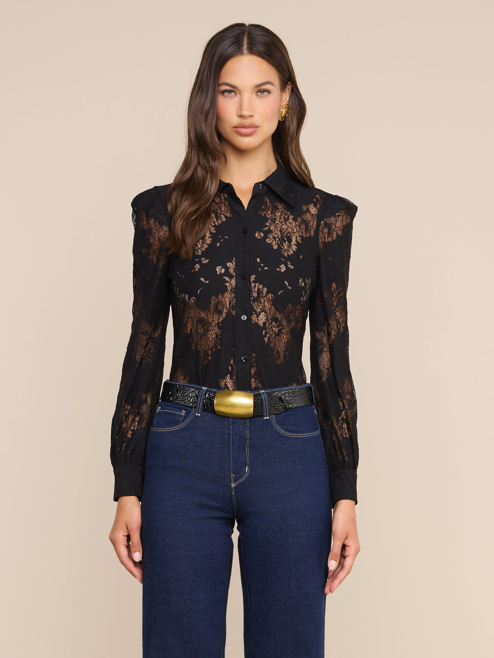 Una donna dai lunghi capelli castani indossa la camicetta in pizzo Jenica, abbinata a jeans blu a vita alta, una cintura nera con fibbia dorata e orecchini dorati, in posa su uno sfondo beige neutro.