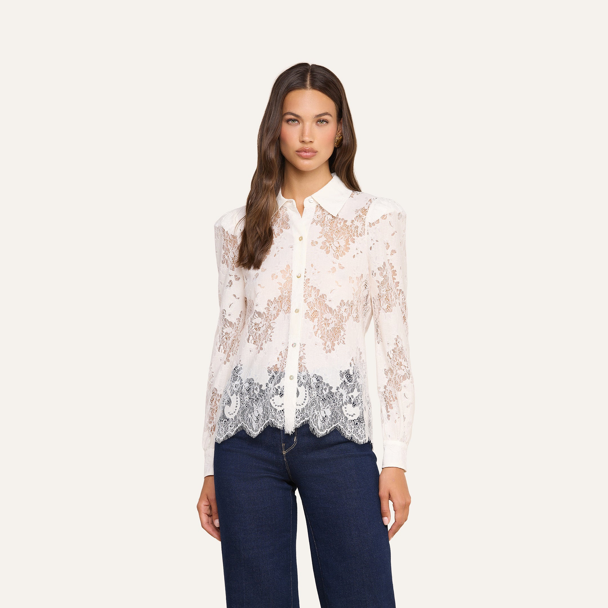 A woman with long brown hair is wearing the Jenica Lace Blouse, a sheer floral lace button-up top with a collar, paired with dark blue jeans and standing against a plain light background.