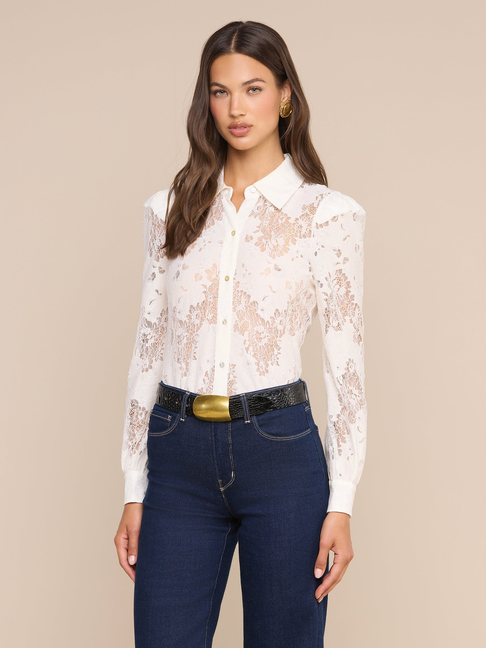 Una donna dai lunghi capelli castani indossa la camicetta Jenica Lace — bianca con pizzo floreale e spalle a sbuffo — abbinata a jeans blu scuro, una cintura nera con una grande fibbia dorata e orecchini a cerchio dorati, in piedi su uno sfondo beige.