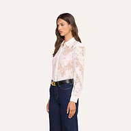 Woman standing sideways wearing the Jenica Lace Blouse—a white, long-sleeve cotton-blend floral lace top with puff shoulders—paired with dark blue pants, a black belt featuring a gold buckle, and gold hoop earrings against a plain light background.