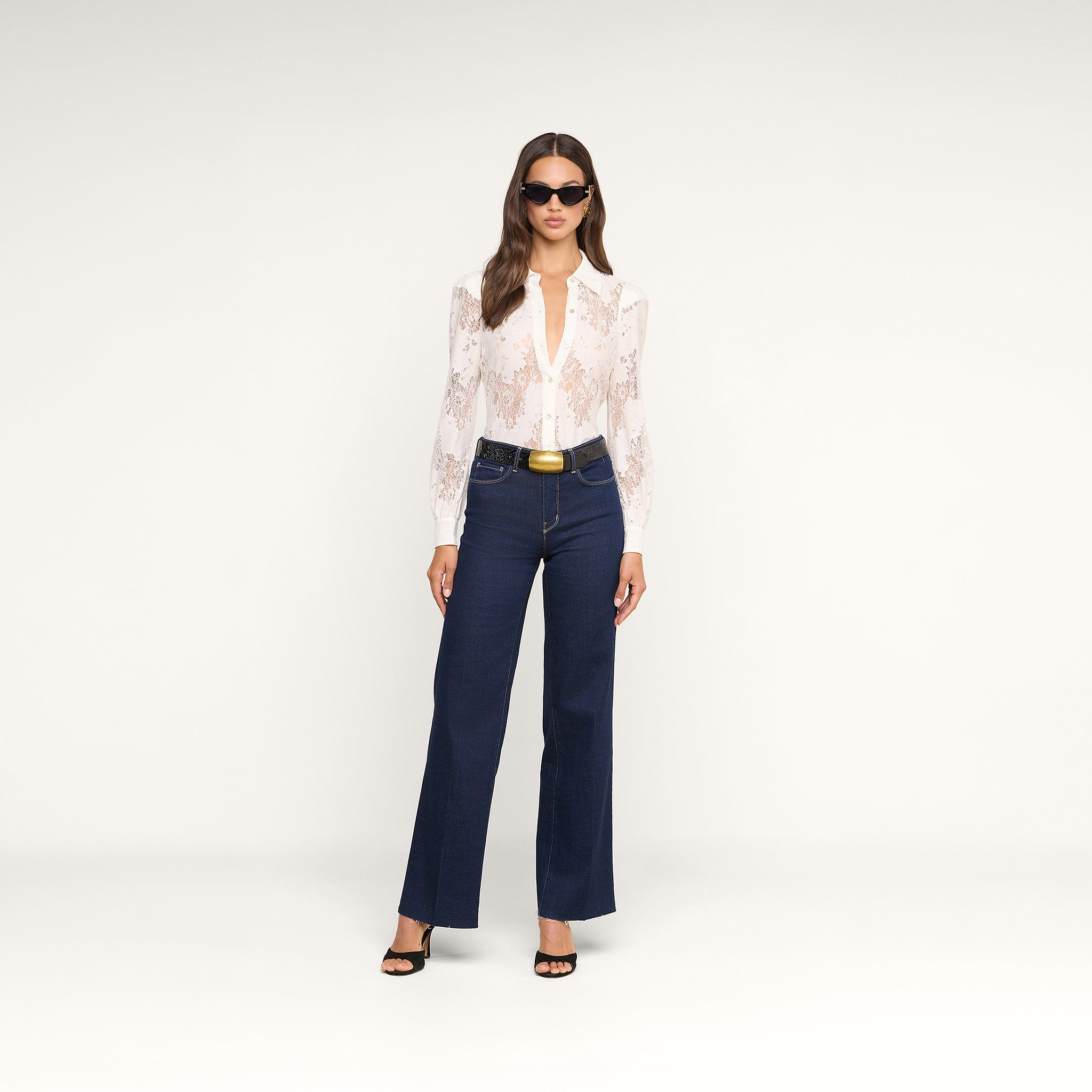 Woman standing against a plain background, wearing sunglasses, the Jenica Lace Blouse with puff shoulders, dark blue high-waisted flared jeans with a black belt, and black heeled sandals.