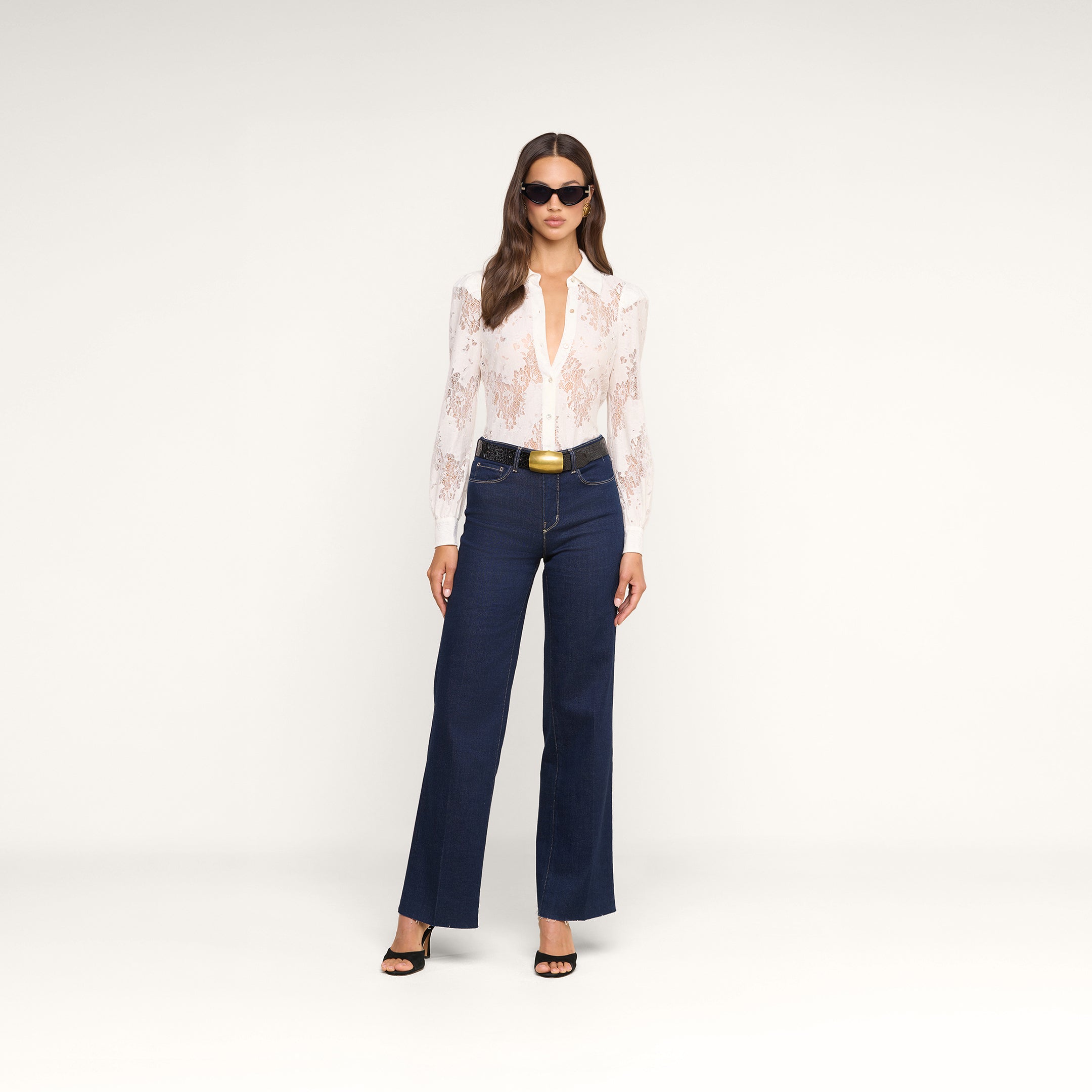 Woman standing against a plain background, wearing sunglasses, the Jenica Lace Blouse with puff shoulders, dark blue high-waisted flared jeans with a black belt, and black heeled sandals.