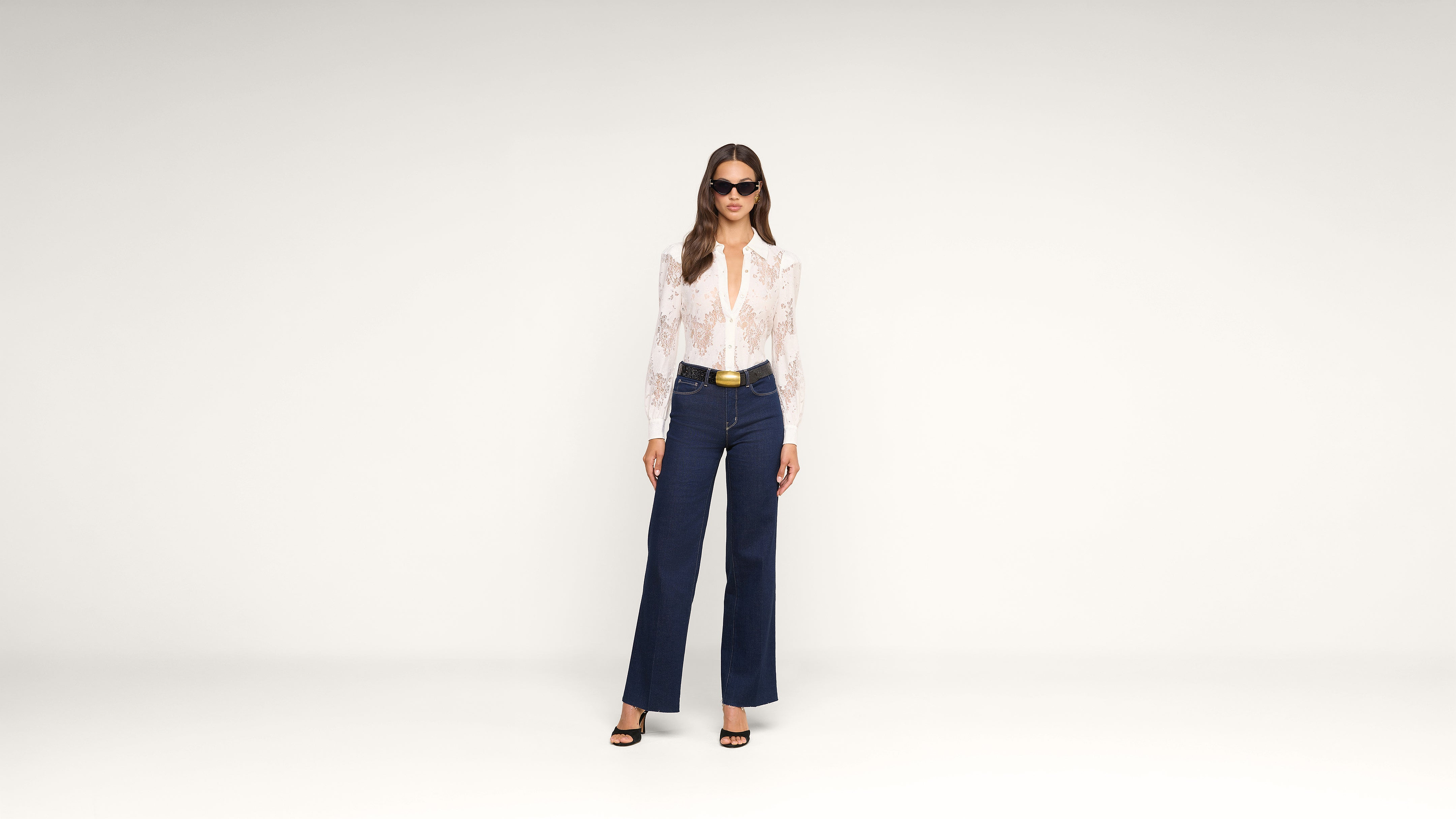 A woman wearing sunglasses, a white lace blouse, dark high-waisted wide-leg jeans, and black heels stands against a plain white background.