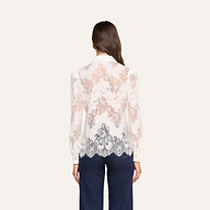 A woman stands with her back to the camera, wearing the Jenica Lace Blouse—a long-sleeve, sheer white floral lace top crafted from a cotton blend. The blouse features intricate patterns, scalloped edges, a collar, and subtle puff shoulders. She pairs it with dark blue pants against a plain, light-colored background.