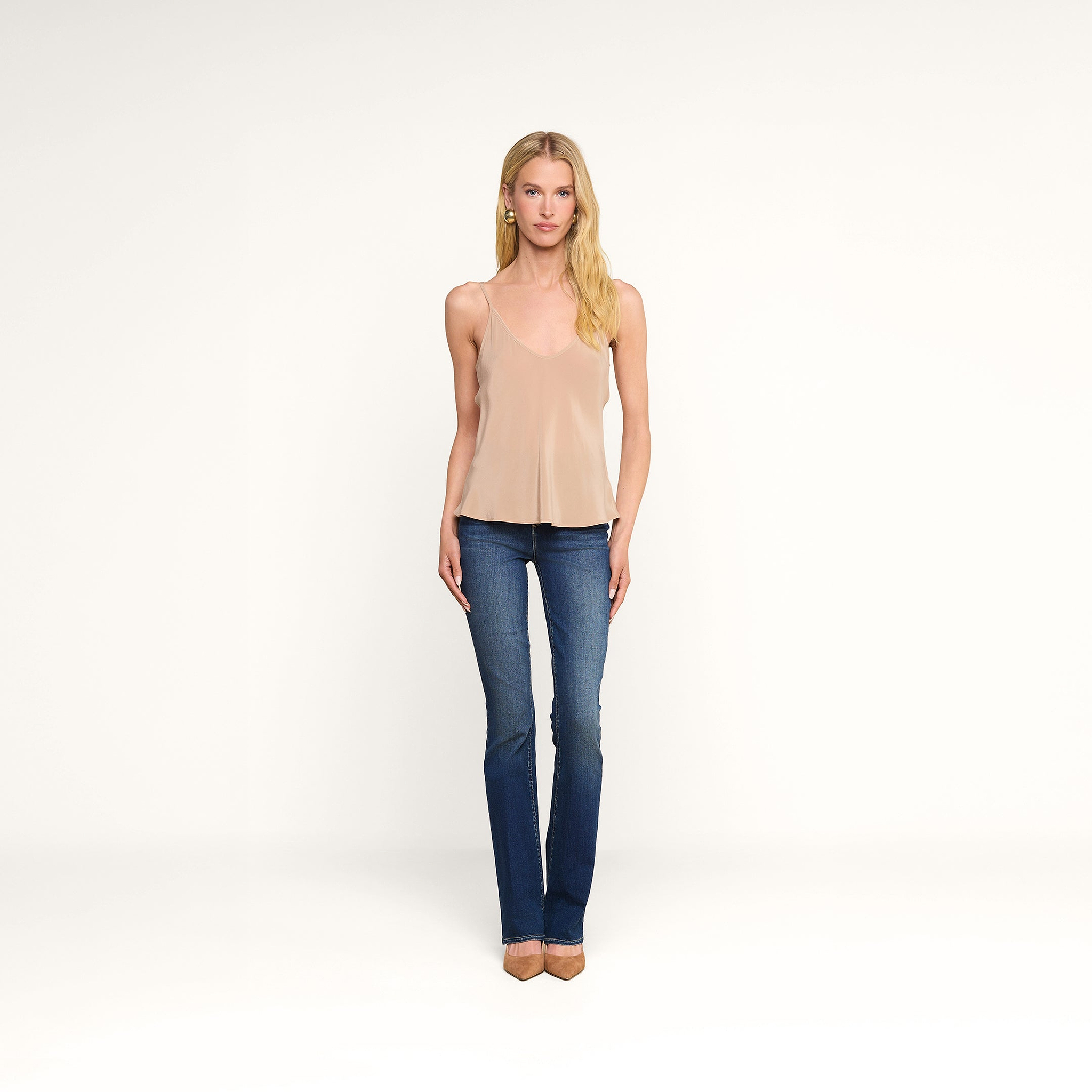 Woman stands facing forward against a plain white background, wearing the Lexi Silk Camisole in beige with adjustable straps, paired with blue jeans, tan heels, and gold earrings. She has long blonde hair and a neutral expression.