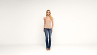 A woman stands facing forward against a plain white background, wearing a beige sleeveless top, blue jeans, and beige shoes. Her arms are relaxed at her sides and she has long, blonde hair.