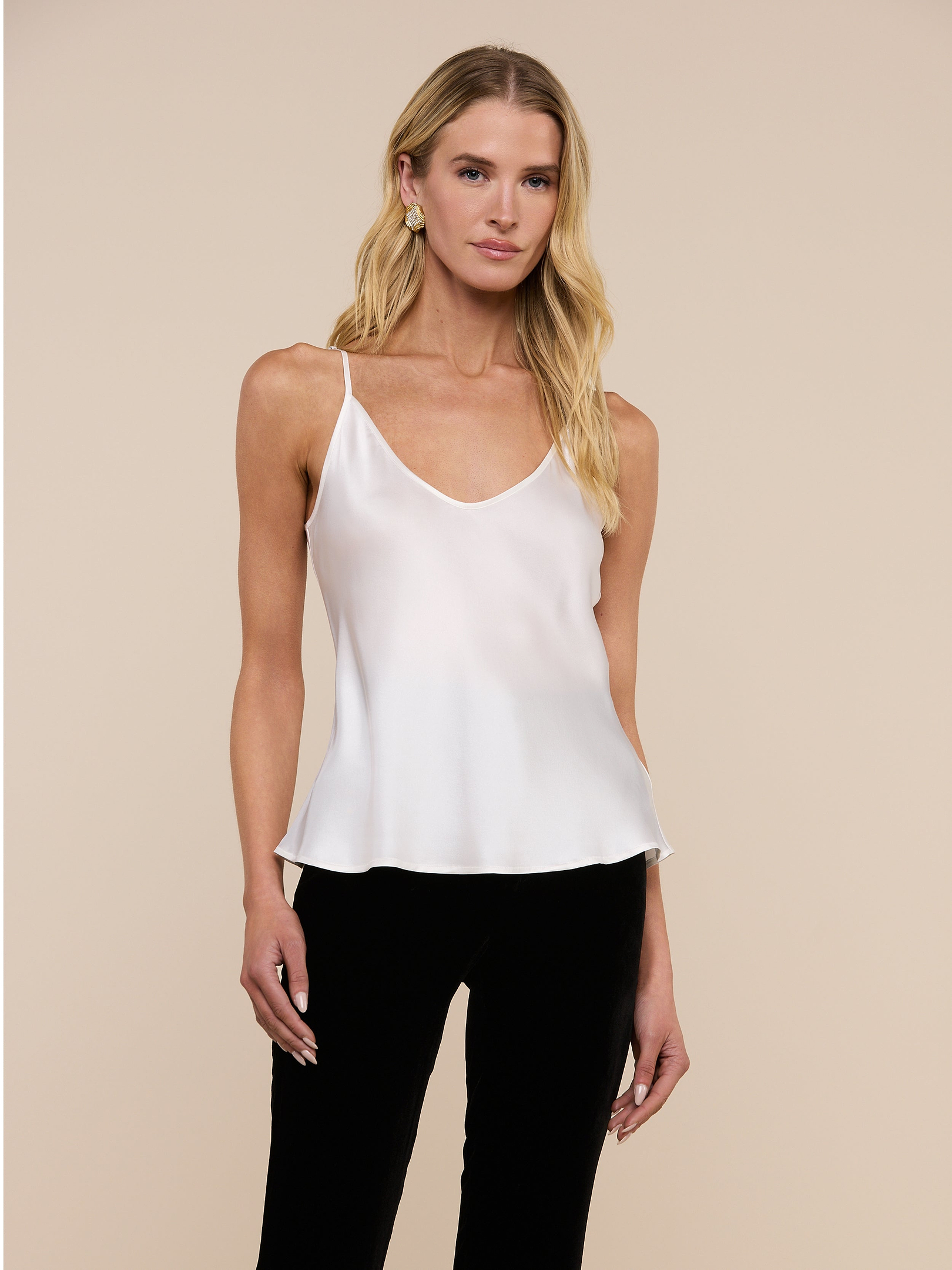 A woman with long blonde hair wears the Lexi Silk Camisole in white, featuring adjustable straps, paired with black pants while standing against a plain light background.