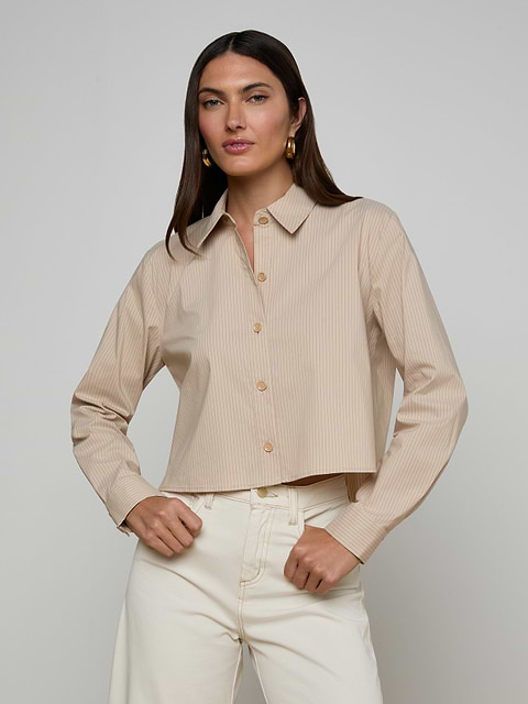 A woman stands with her hands on her hips, wearing the Cosette Shirt—a tailored, beige long-sleeve button-up—paired with off-white pants. She has long, straight brown hair and faces forward against a plain light background.