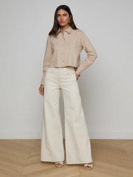 A woman stands against a plain white background, wearing the Cosette Shirt made from organic cotton with long sleeves, paired with high-waisted, wide-leg white pants. She has long dark hair and is wearing open-toed heels.