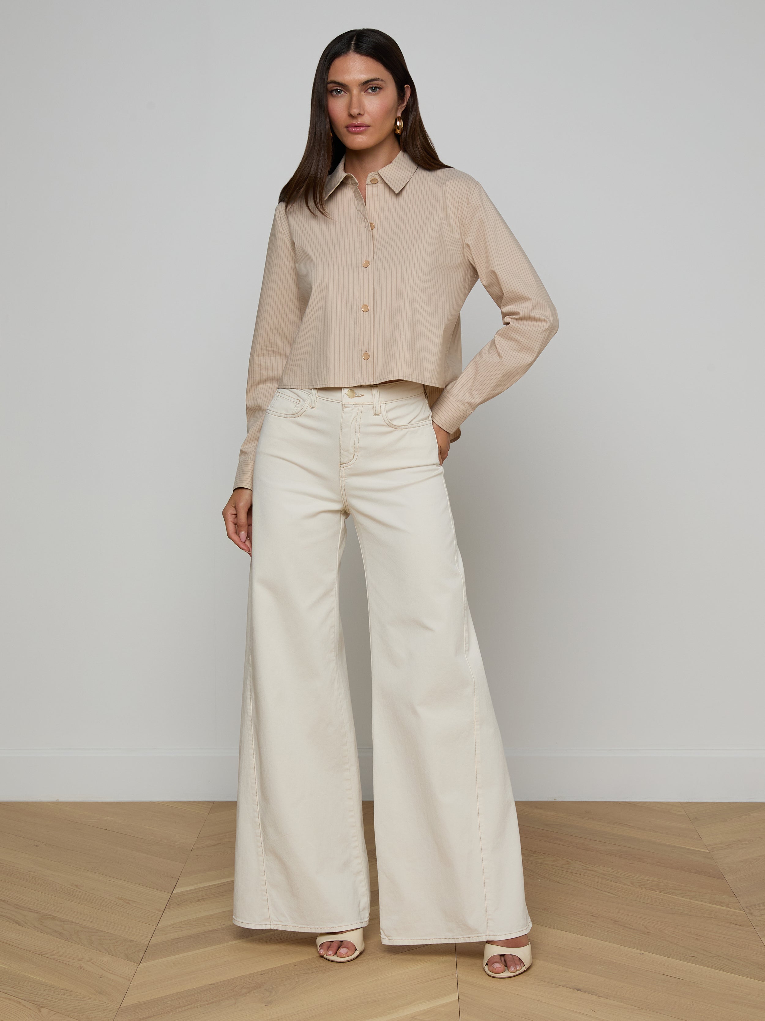 A woman stands against a plain white background, wearing the Cosette Shirt made from organic cotton with long sleeves, paired with high-waisted, wide-leg white pants. She has long dark hair and is wearing open-toed heels.