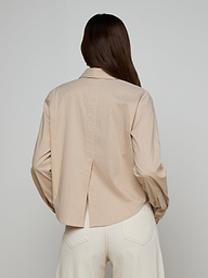 A person with long brown hair, seen from the back, wears the Cosette Shirt—a beige, collared, tailored shirt with long sleeves and a back slit—paired with white pants. The background is plain and light-colored.