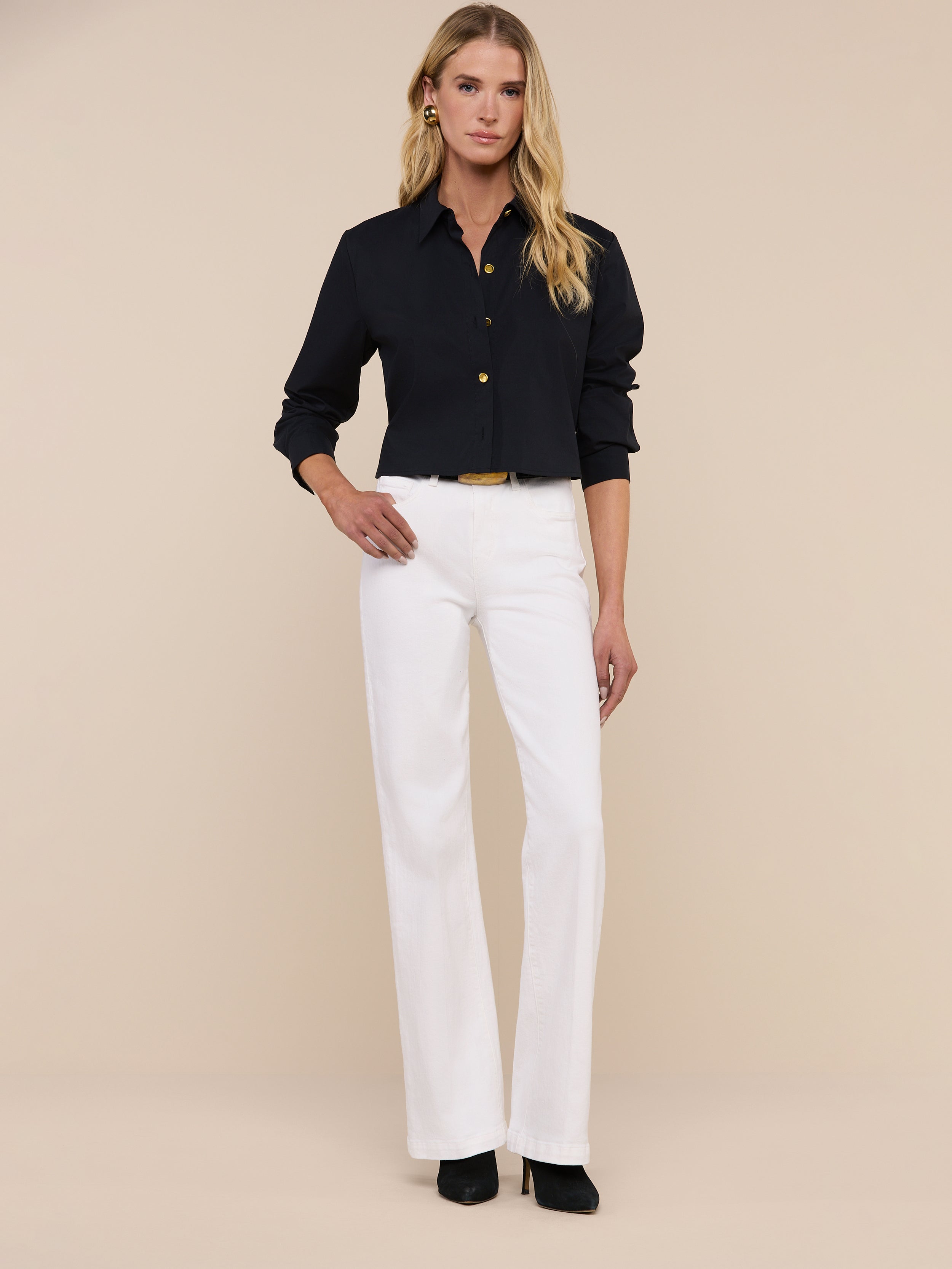 A woman with long blonde hair wears sunglasses, the Cosette Shirt—a tailored black button-up—blue jeans, and black heels as she stands against a plain white background.