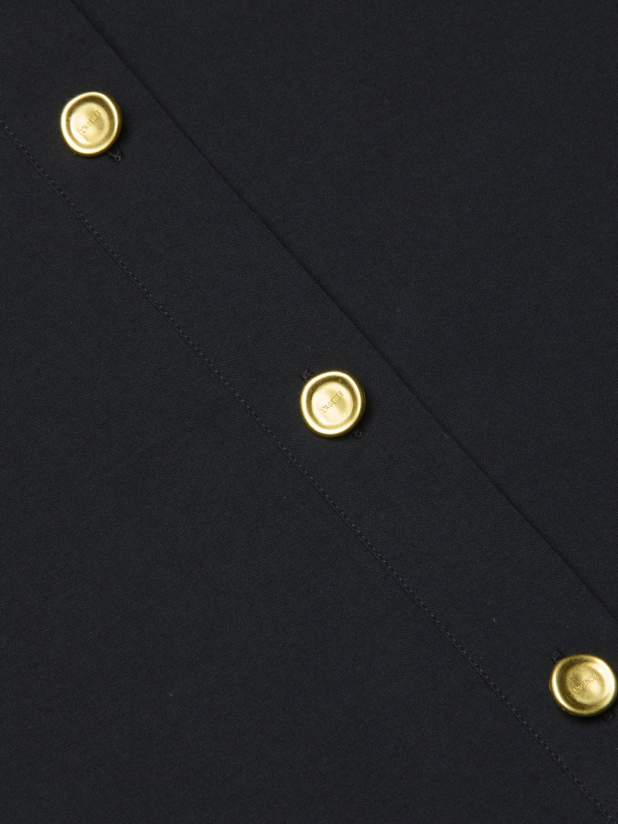 Close-up of the Cosette Shirt in black organic cotton fabric, featuring a row of shiny gold round buttons evenly spaced along a stitched seam.