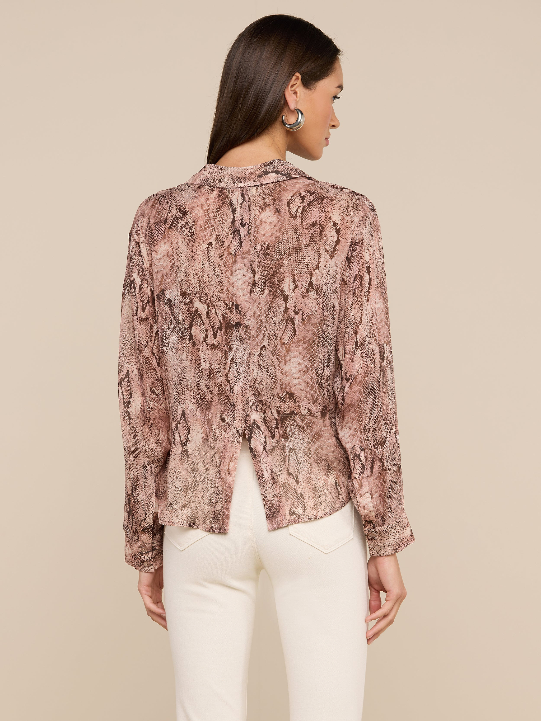 A woman with long brown hair, dressed in the Cosette Shirt—a lightweight button-down featuring a snake print and back slit—paired with white pants, stands against a light background with her back turned.