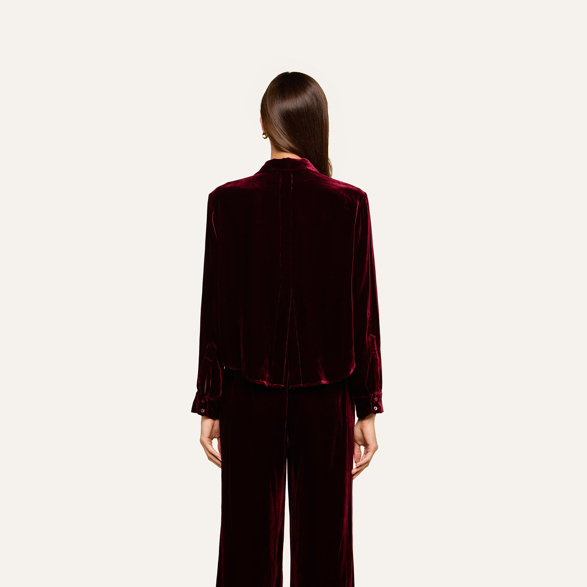 A person with long brown hair, dressed in the Cosette Velvet Shirt in burgundy with matching pants, stands facing away against a plain, light background.
