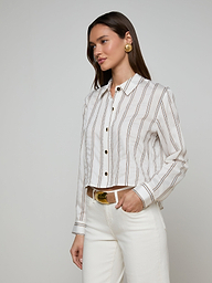 A woman wears the Cosette Shirt—a white button-up tailored shirt with brown jacquard stripes—paired with white pants, a tan belt, and large round earrings, standing against a plain light background.
