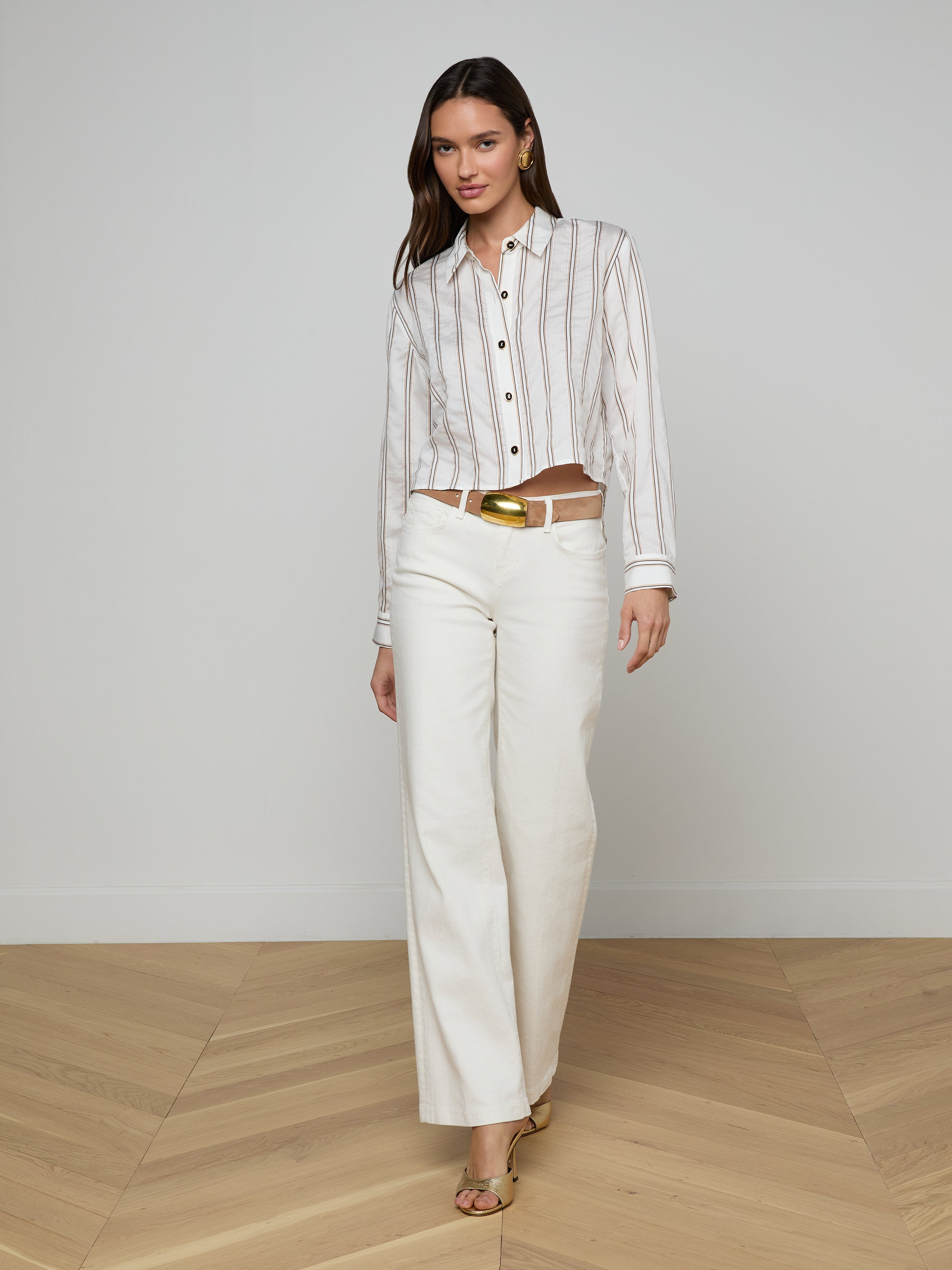 A woman wearing the Cosette Shirt with jacquard stripes, white wide-leg pants, a gold belt, and sandals stands against a plain white background. She has straight dark hair.